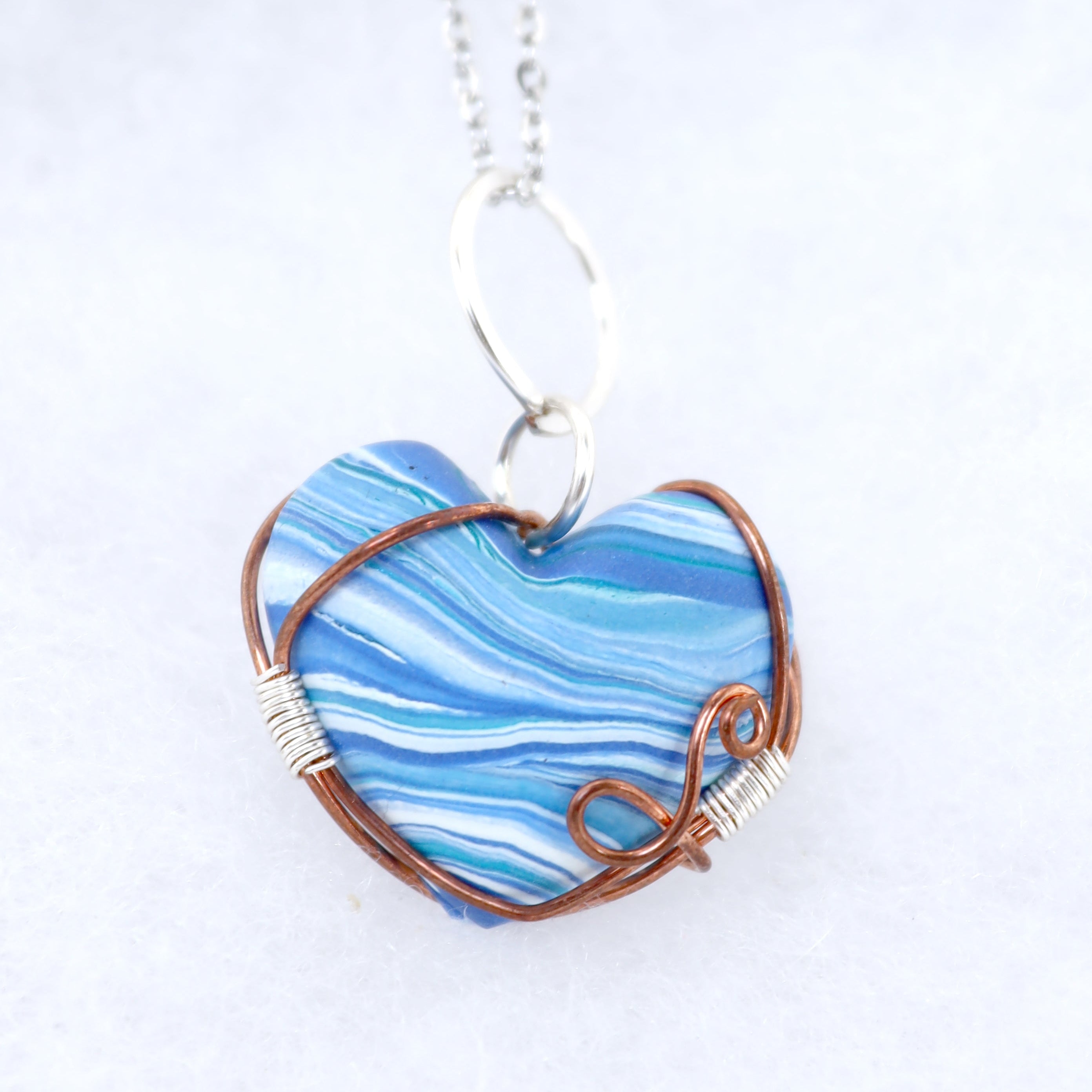 A heart-shaped pendant necklace featuring a blue and white marbled clay center, wrapped in copper wire with silver wire accents, hanging from a silver chain on a white background.