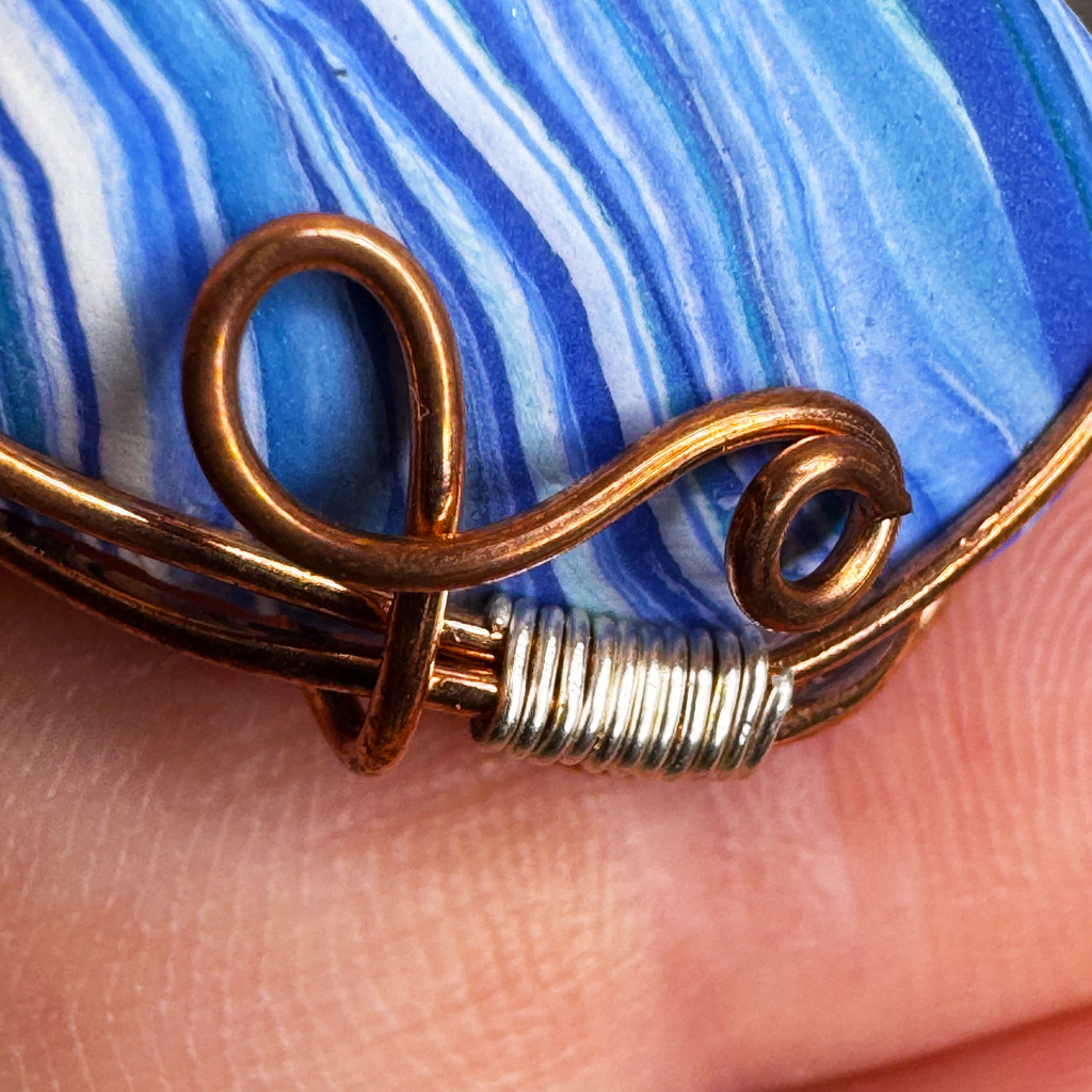 Close-up of a handmade wire-wrapped pendant featuring blue and white marbled clay, wrapped in copper-colored wire with a silver-colored coil detail, held against skin.