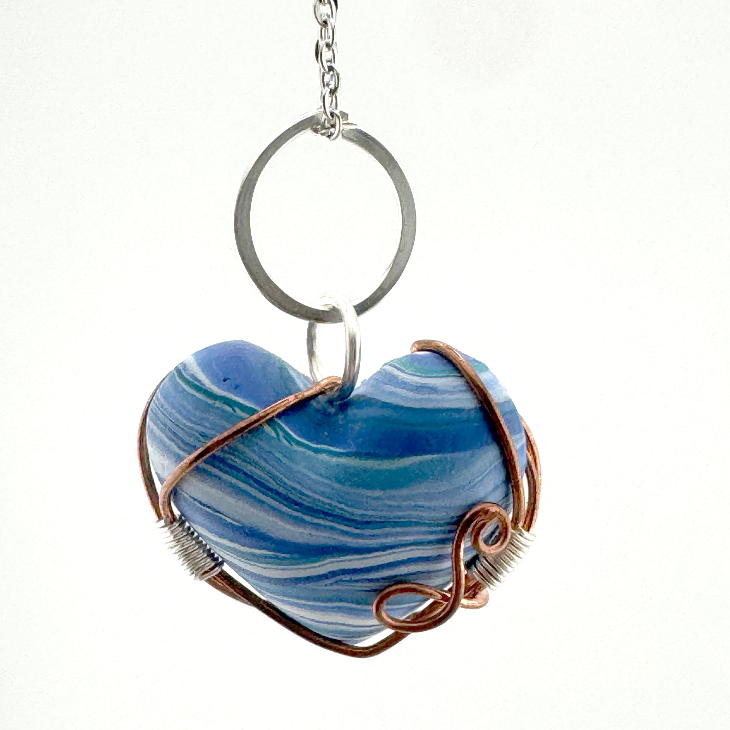 A heart-shaped pendant made of blue and white marbled clay, wrapped in copper wire, hanging from a silver metal ring on a chain against a white background.