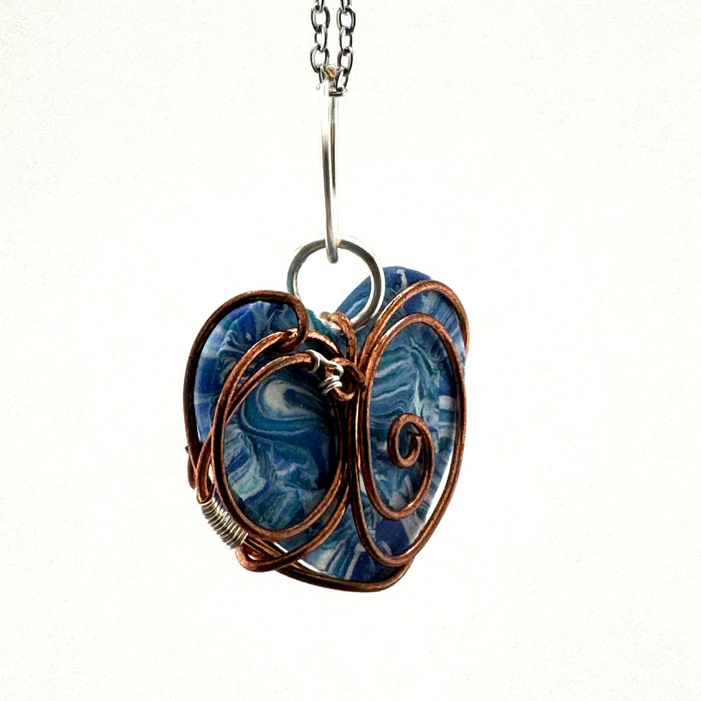A handmade heart-shaped pendant necklace featuring a blue and white marbled clay piece wrapped in copper wire, suspended from a silver-toned chain against a plain white background.