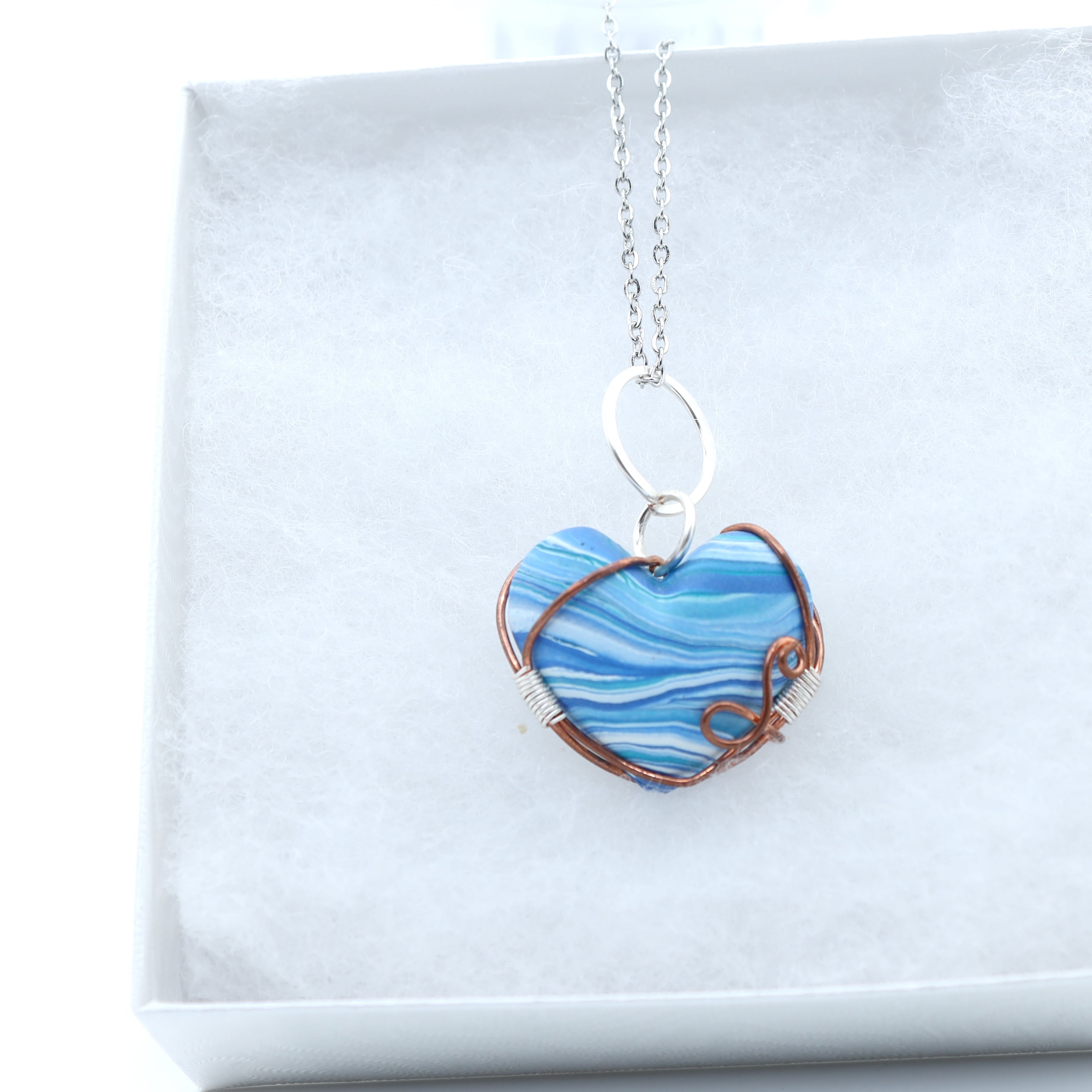 A heart-shaped pendant necklace with blue and white marbled clay, wrapped in copper wire, hanging from a silver chain in a white display box.