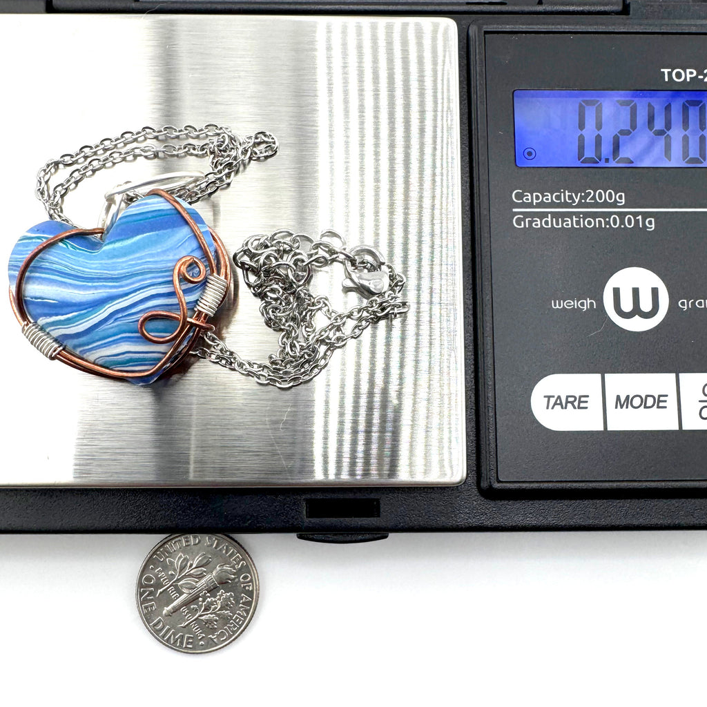 A silver chain necklace with a heart-shaped marbled blue and white clay pendant featuring a copper-toned border, shown resting on a digital scale. 