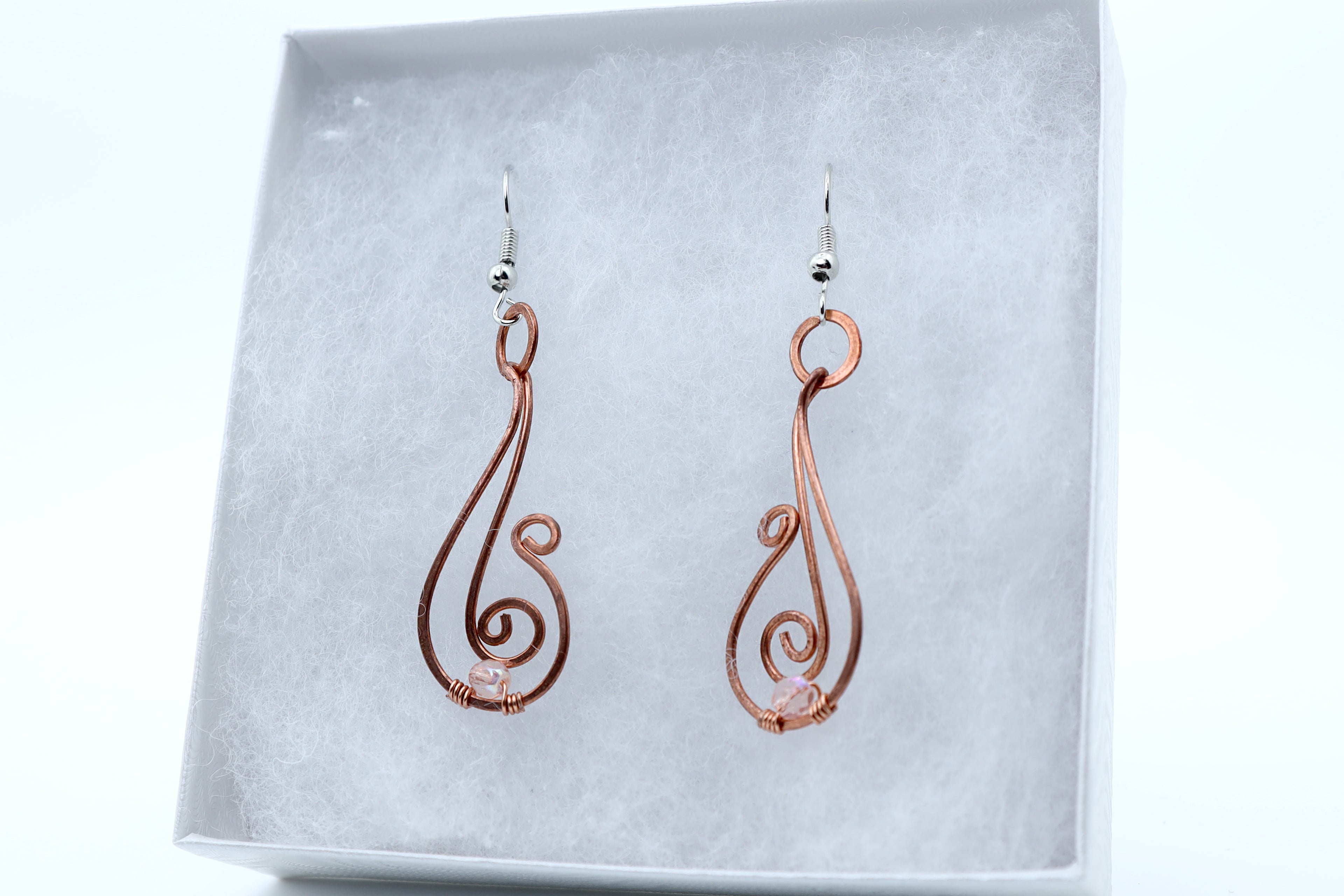 A pair of copper wire dangle earrings with silver fishhook backings, displayed inside a white jewelry box with a textured gray insert.