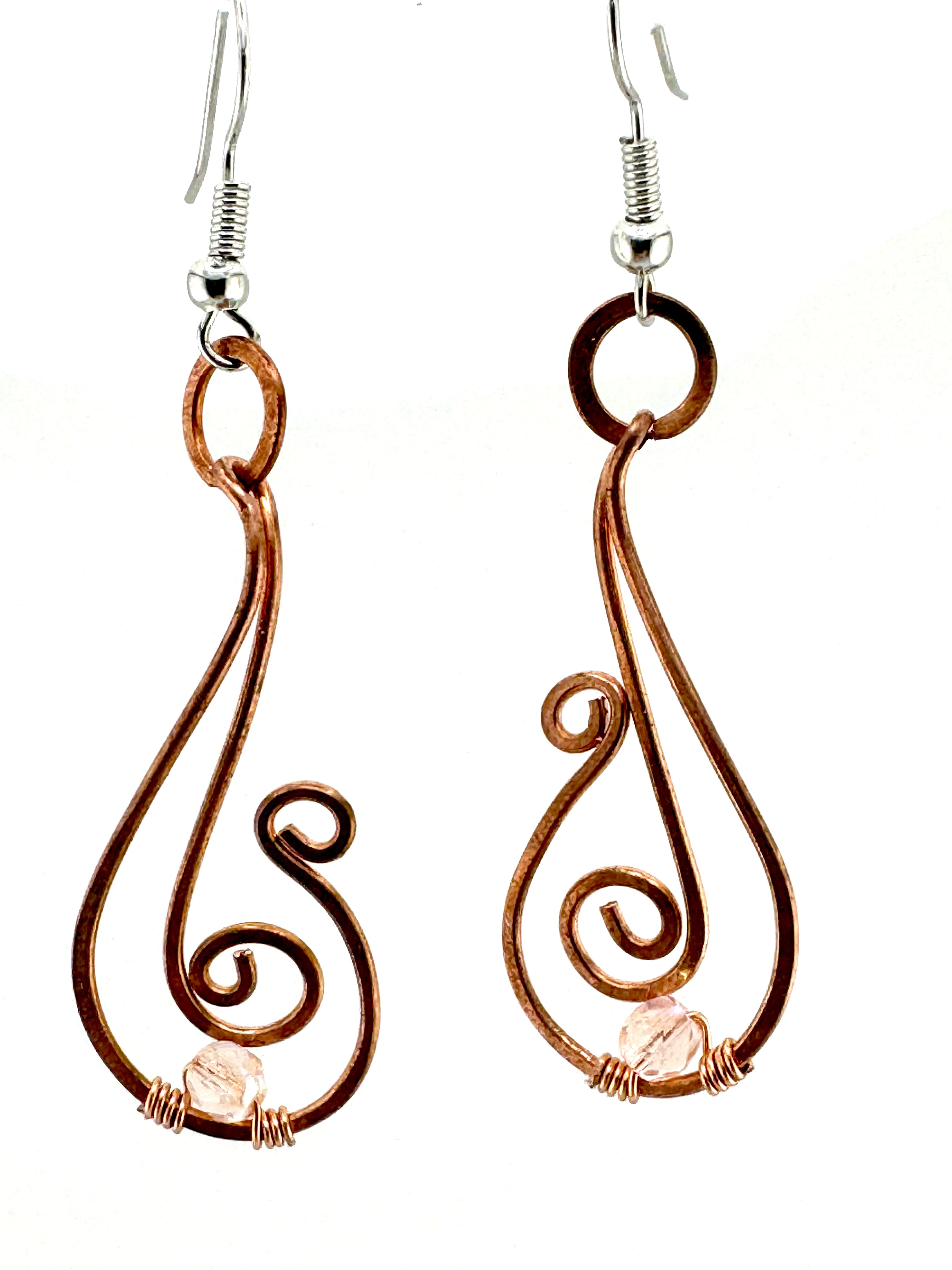 A pair of handmade wire earrings with copper-colored loops and swirls, featuring small pale pink gemstone accents and silver fishhook ear wires, shown against a plain white background.