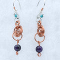 A pair of handmade dangle earrings featuring copper wire coils, silver hooks, small turquoise beads, and dark purple round beads, displayed on a textured light background.