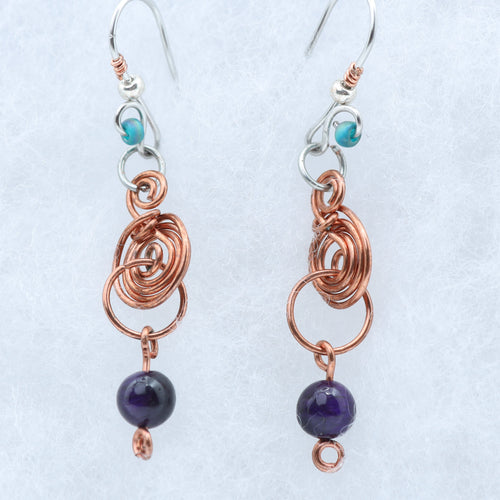 A pair of handmade dangle earrings featuring copper wire coils, silver hooks, small turquoise beads, and dark purple round beads, displayed on a textured light background.