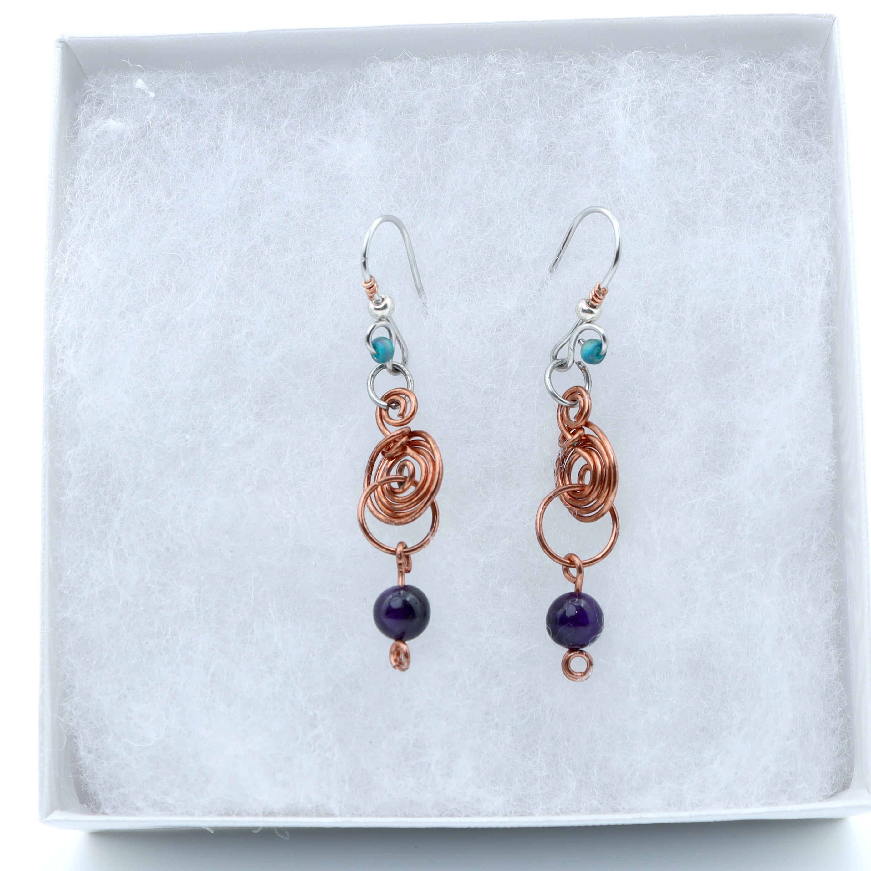 A pair of handmade dangle earrings displayed on a white textured jewelry box. The earrings feature copper-colored coiled wire, small turquoise blue beads, and dark purple round beads hanging from silver fishhook wires.