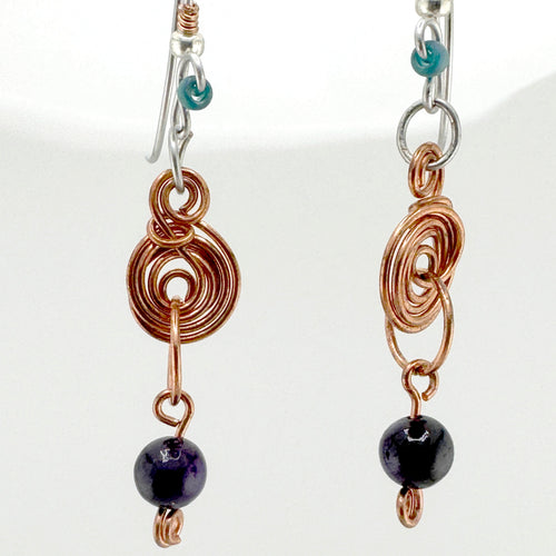 A pair of handmade dangle earrings featuring copper-colored coiled wire, a small turquoise bead, and a dark purple round bead at the bottom, shown against a plain white background.