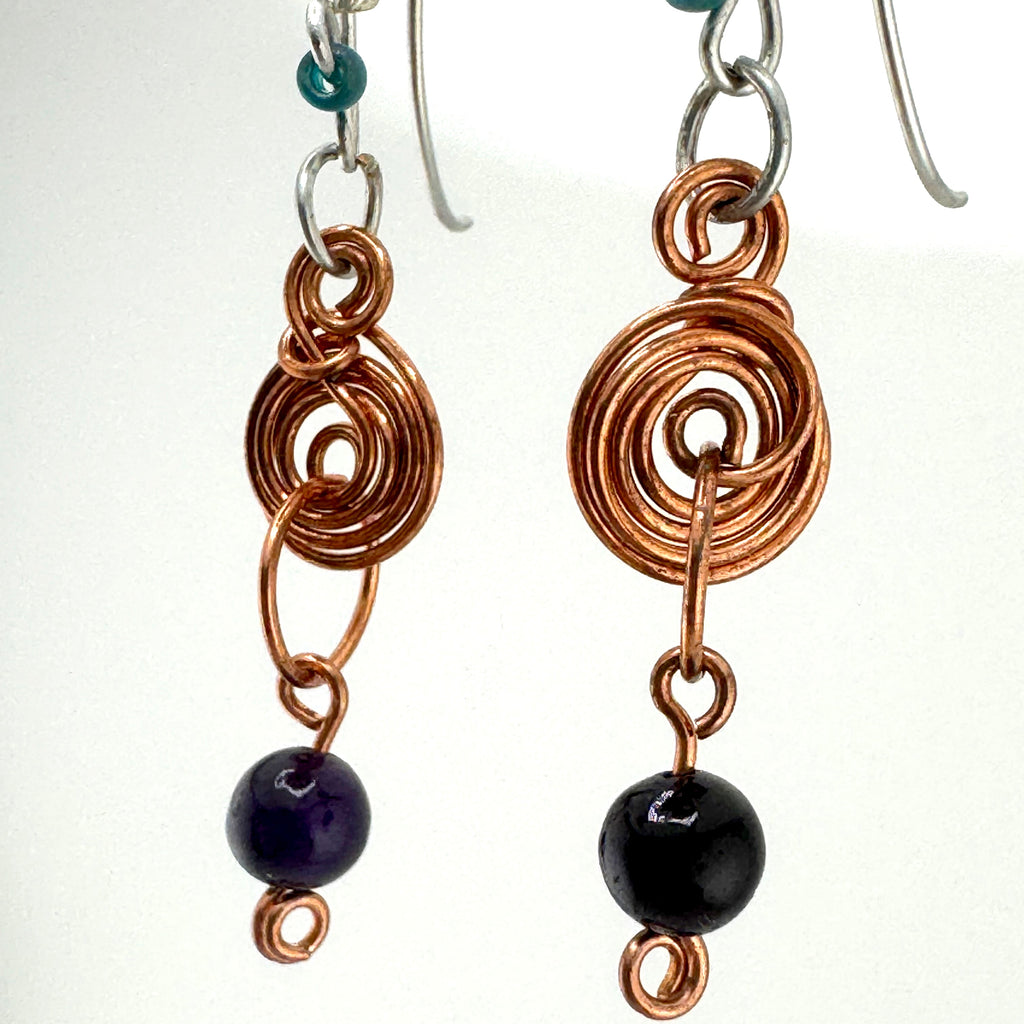 A pair of handmade dangle earrings featuring copper wire spirals, silver fishhook wires, a small teal bead, and a dark purple bead at the bottom.