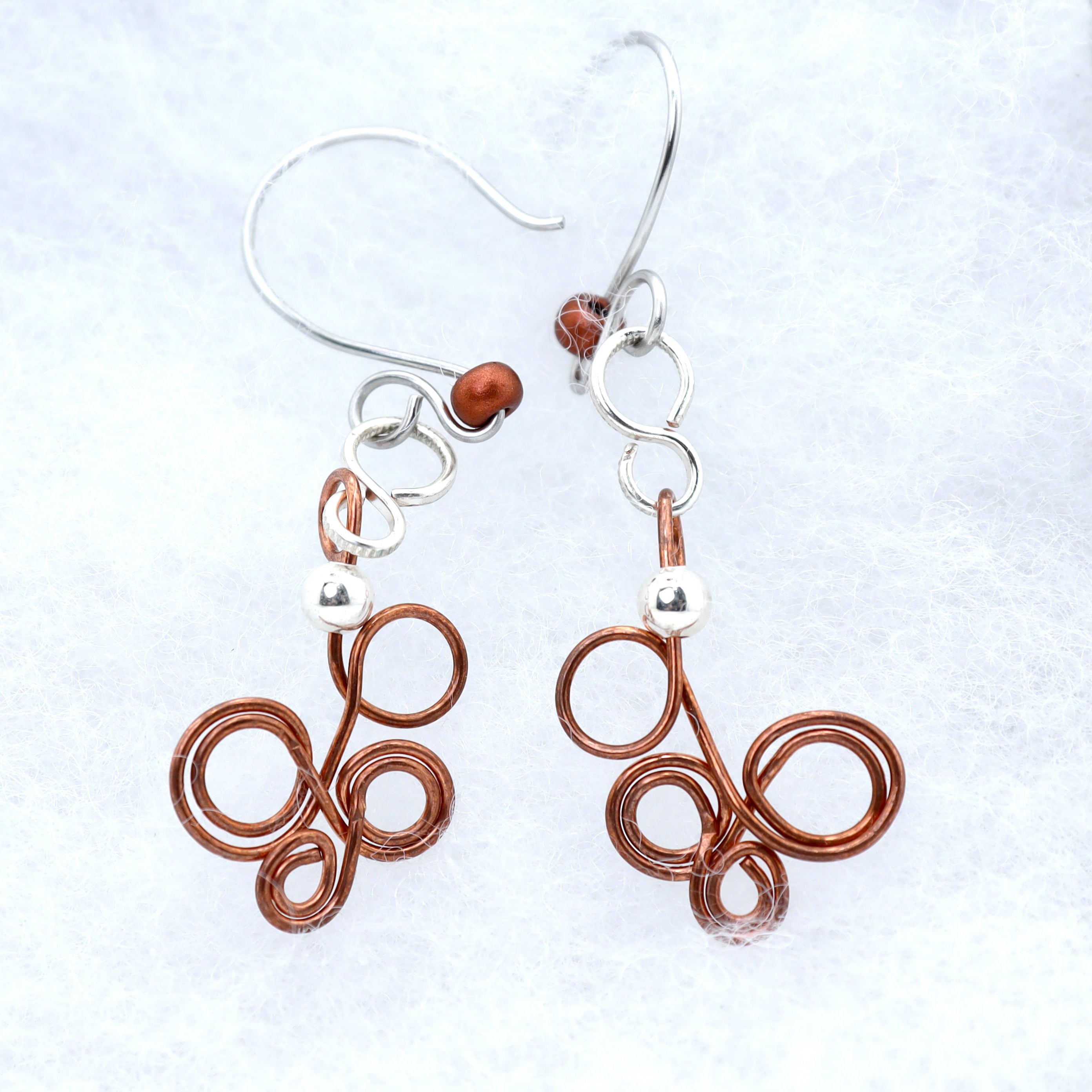 A pair of handmade dangle earrings featuring copper-colored coiled wire and silver fishhook wires, displayed on a textured white background.