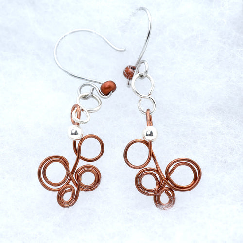 A pair of handmade dangle earrings featuring copper-colored coiled wire and silver fishhook wires, displayed on a textured white background.