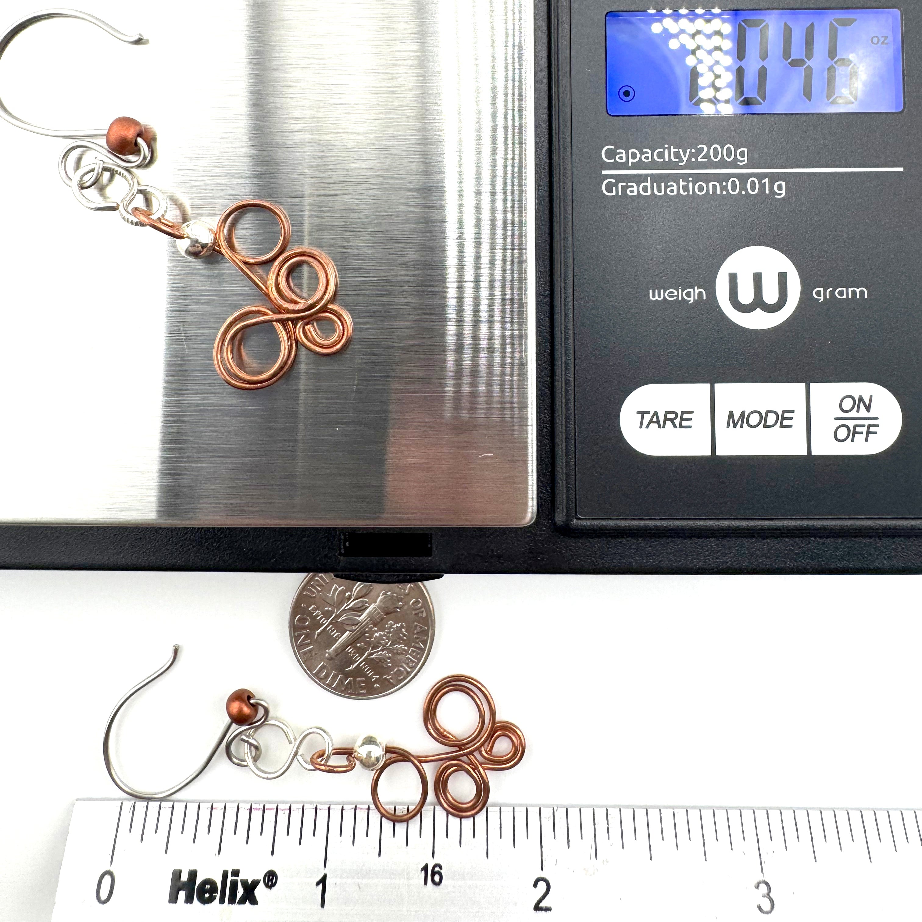 A pair of handmade copper wire earrings with silver hooks and small copper beads, shown alongside a digital scale and a Helix brand ruler for size reference.