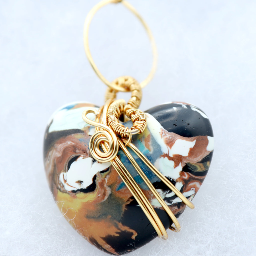 A heart-shaped pendant with a marbled pattern of black, brown, white, and teal, wrapped in gold wire with a gold hanging loop, shown on a light gray background.