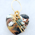 A heart-shaped pendant with a marbled pattern of black, brown, white, and teal, wrapped in gold wire with a gold hanging loop, shown on a light gray background.