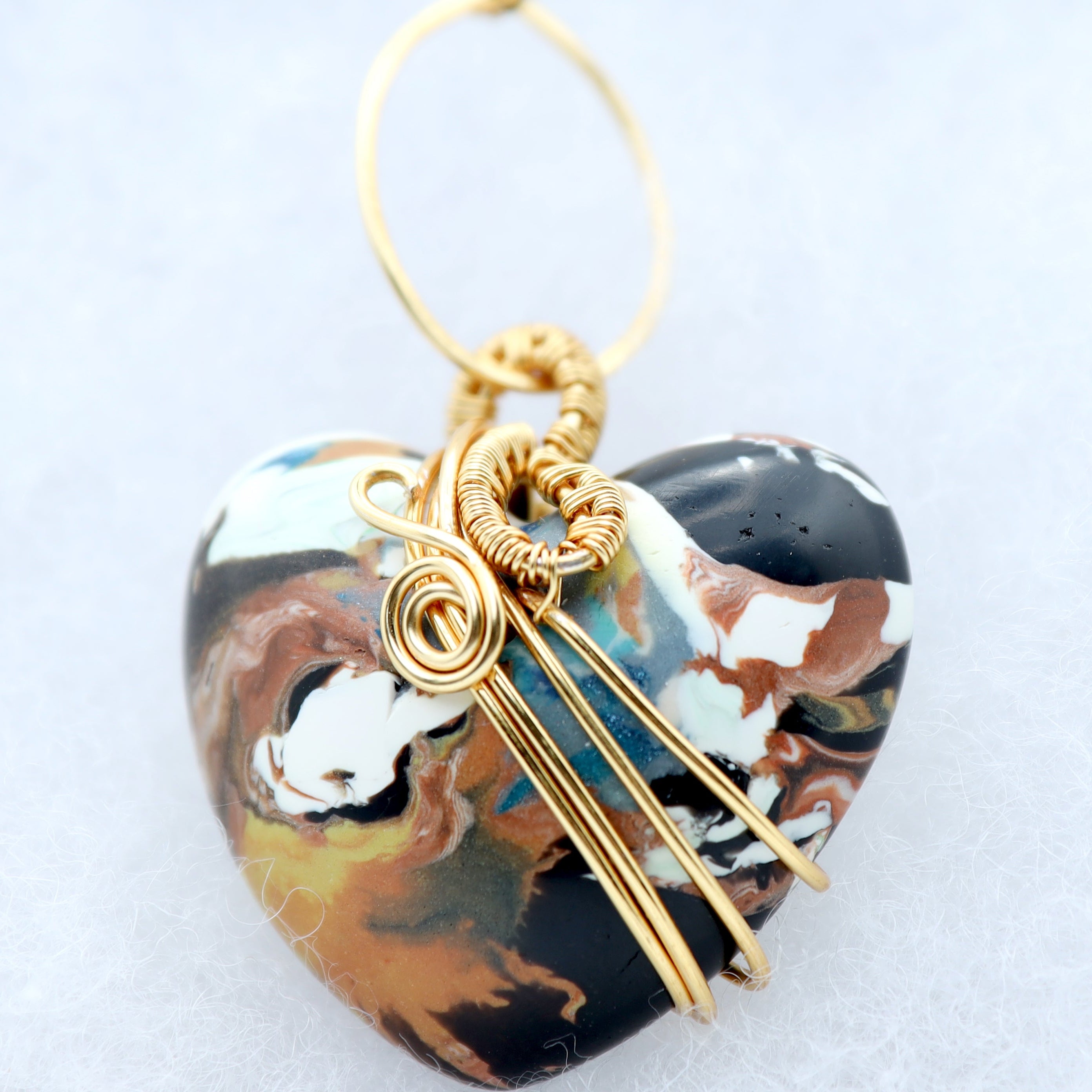 A heart-shaped pendant with a marbled pattern of black, brown, white, and teal, wrapped in gold wire with a gold hanging loop, shown on a light gray background.