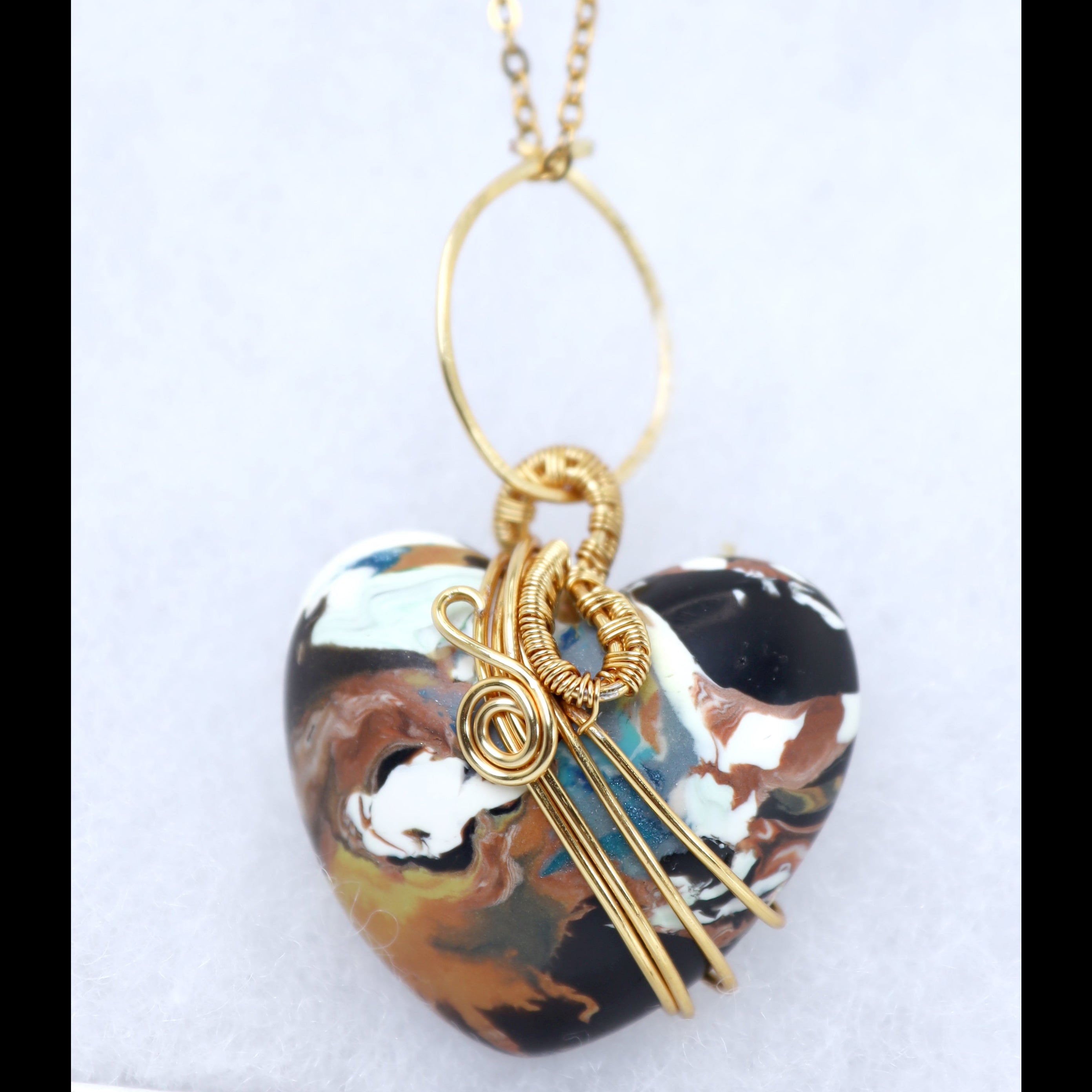 A gold chain necklace with a heart-shaped pendant featuring a marbled pattern of black, brown, white, and light blue. The pendant is wrapped with gold wire loops and bars, photographed against a light gray background.
