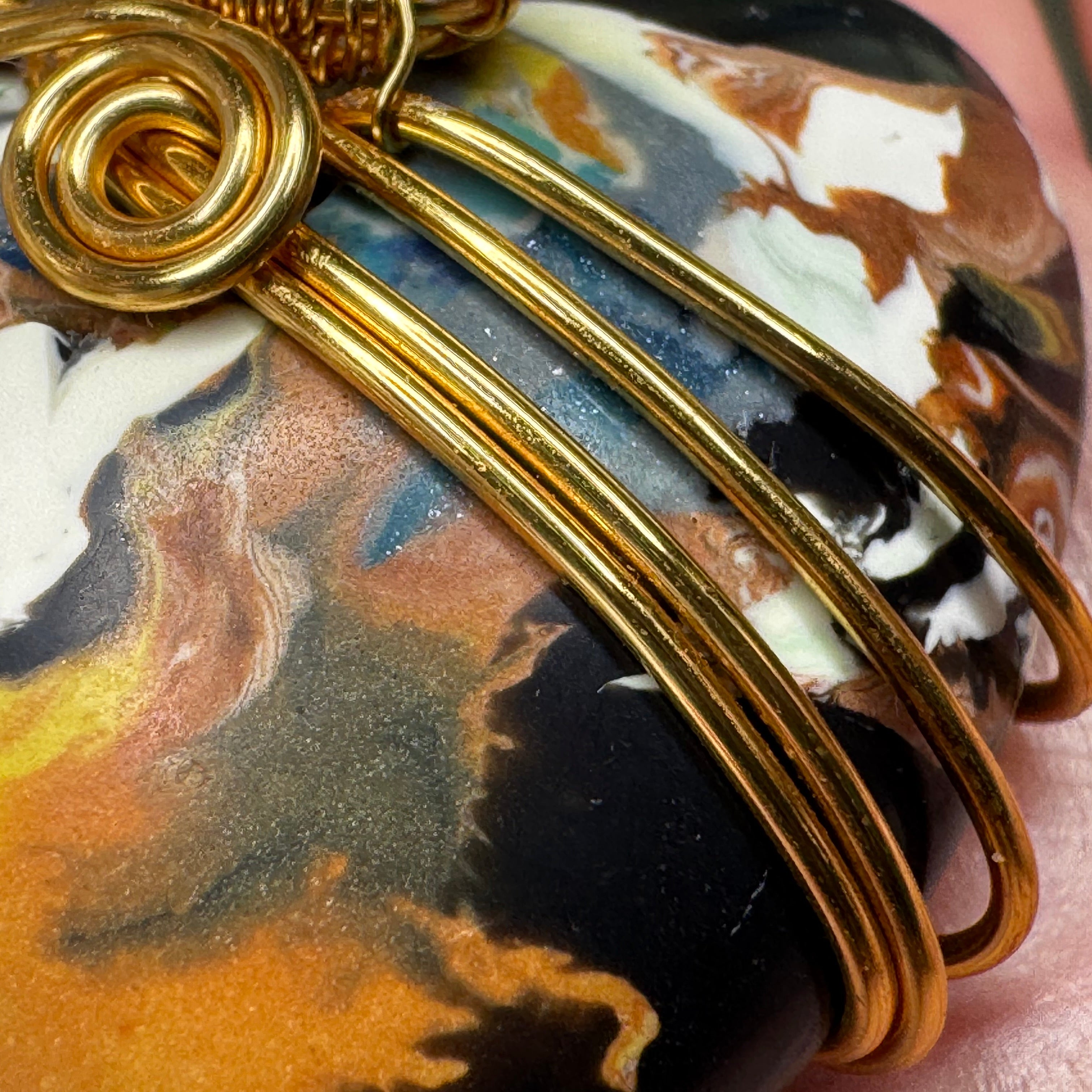 Close-up of a gold wire-wrapped clay pendant  featuring black, white, brown, and gold swirls, held against a skin background.