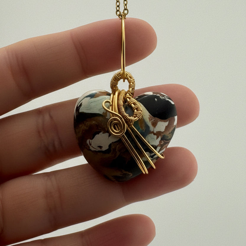 A close-up of a hand holding a polished marbled clay pendant wrapped in gold wire, attached to a gold chain, against a plain light background.