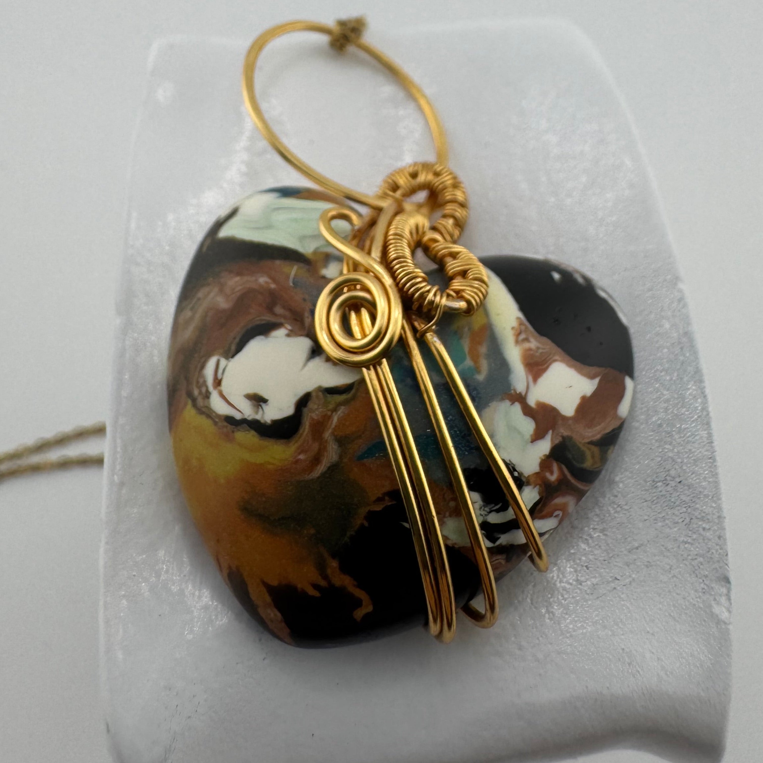 A heart-shaped pendant with a marbled black, brown, and white pattern, wrapped in gold wire with a gold loop, displayed on a white stand.