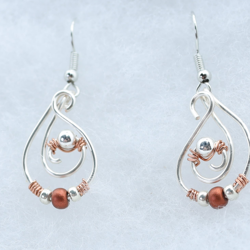 A pair of silver teardrop dangle earrings with copper wire swirls, small silver beads, and a round copper bead, displayed on a light gray textured background.
