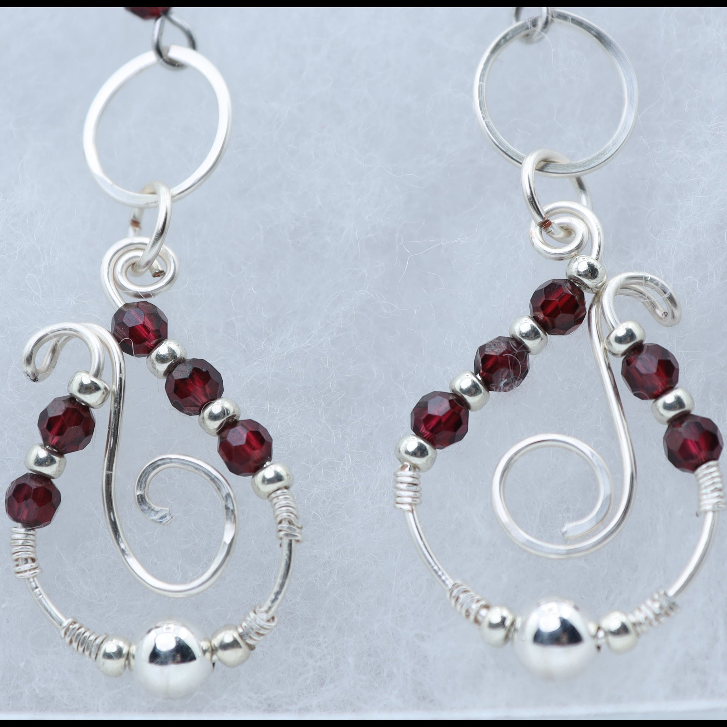 A pair of silver dangle earrings with faceted dark red garnet gemstone and silver wire swirls, displayed on a light gray textured background.