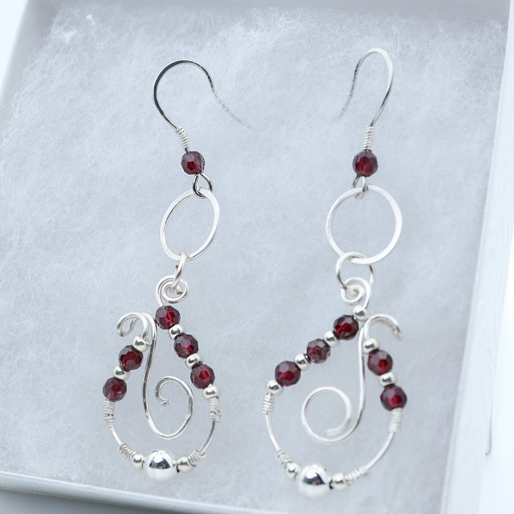 A pair of silver dangle earrings with small dark red garnet accents, displayed inside a white jewelry box.