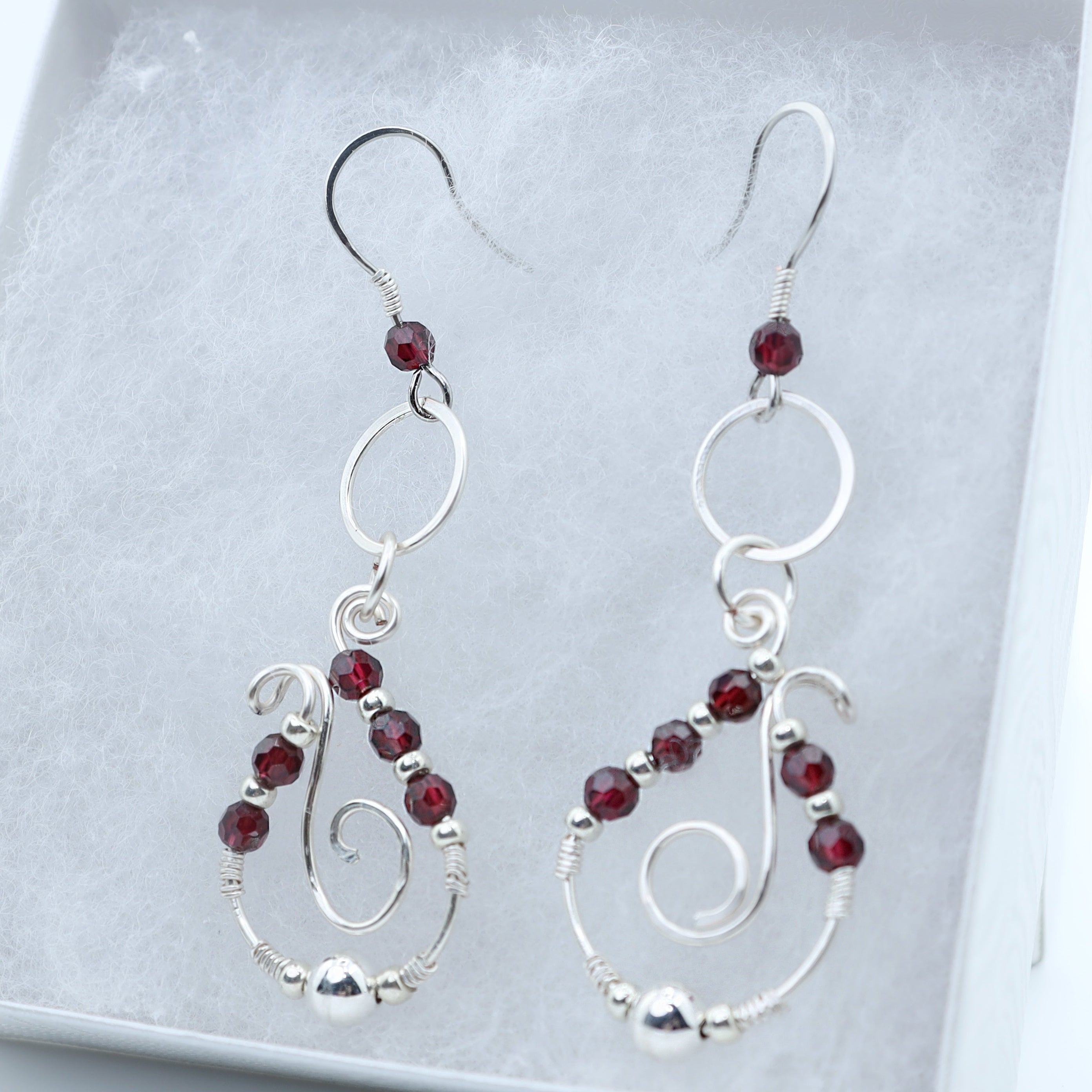 A pair of silver dangle earrings with small dark red garnet accents, displayed inside a white jewelry box.