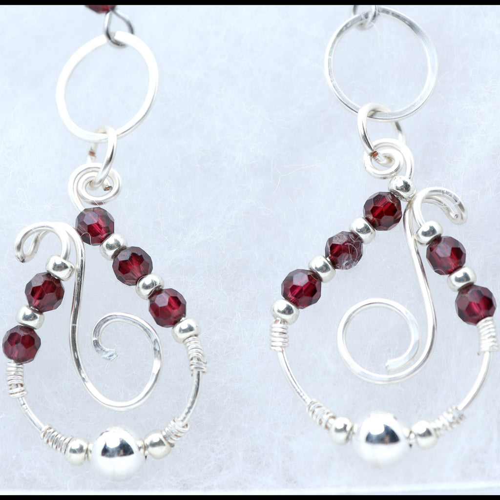 A pair of silver dangle earrings with faceted dark red garnet gemstone beads and silver wire swirls, displayed on a light textured background.
