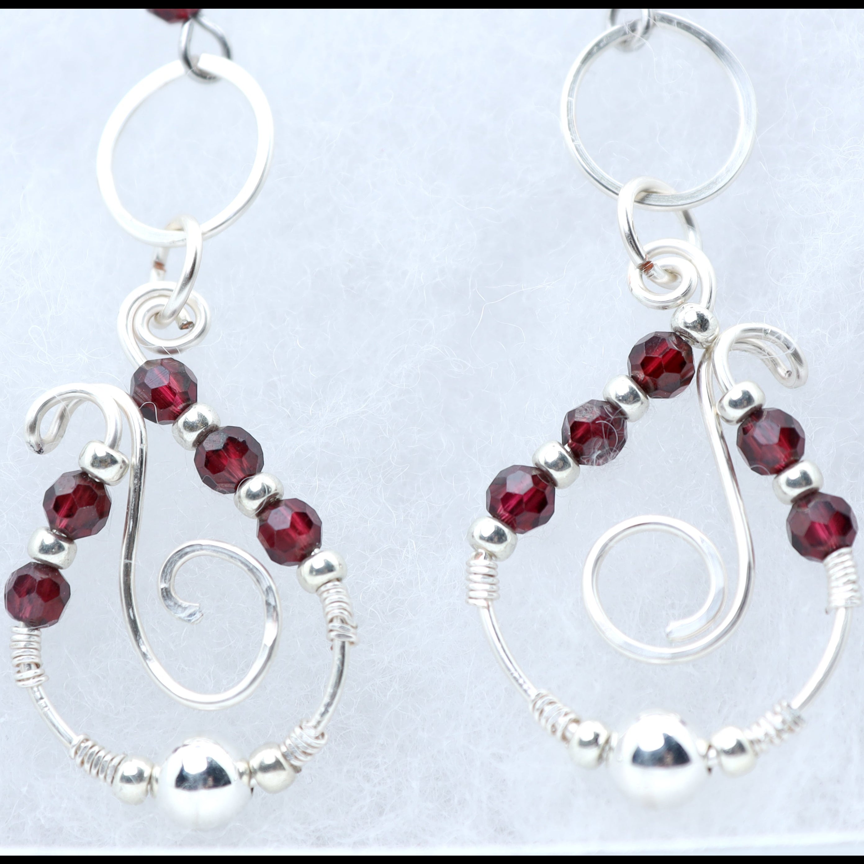 A pair of silver dangle earrings with faceted dark red garnet gemstone beads and silver wire swirls, displayed on a light textured background.