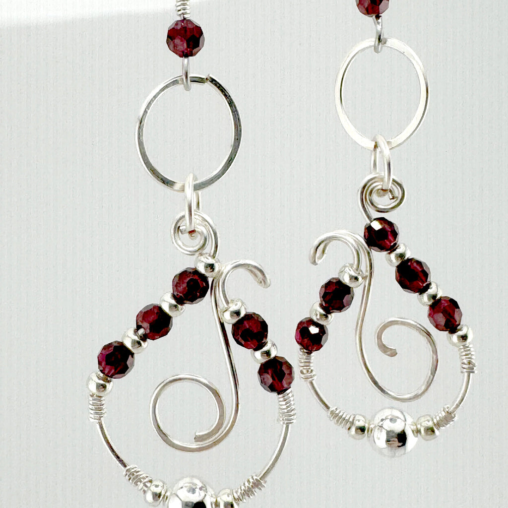 A pair of silver-toned dangle earrings with faceted dark red garnet stones, open circular hoops, and spiral wire details, shown against a light background.