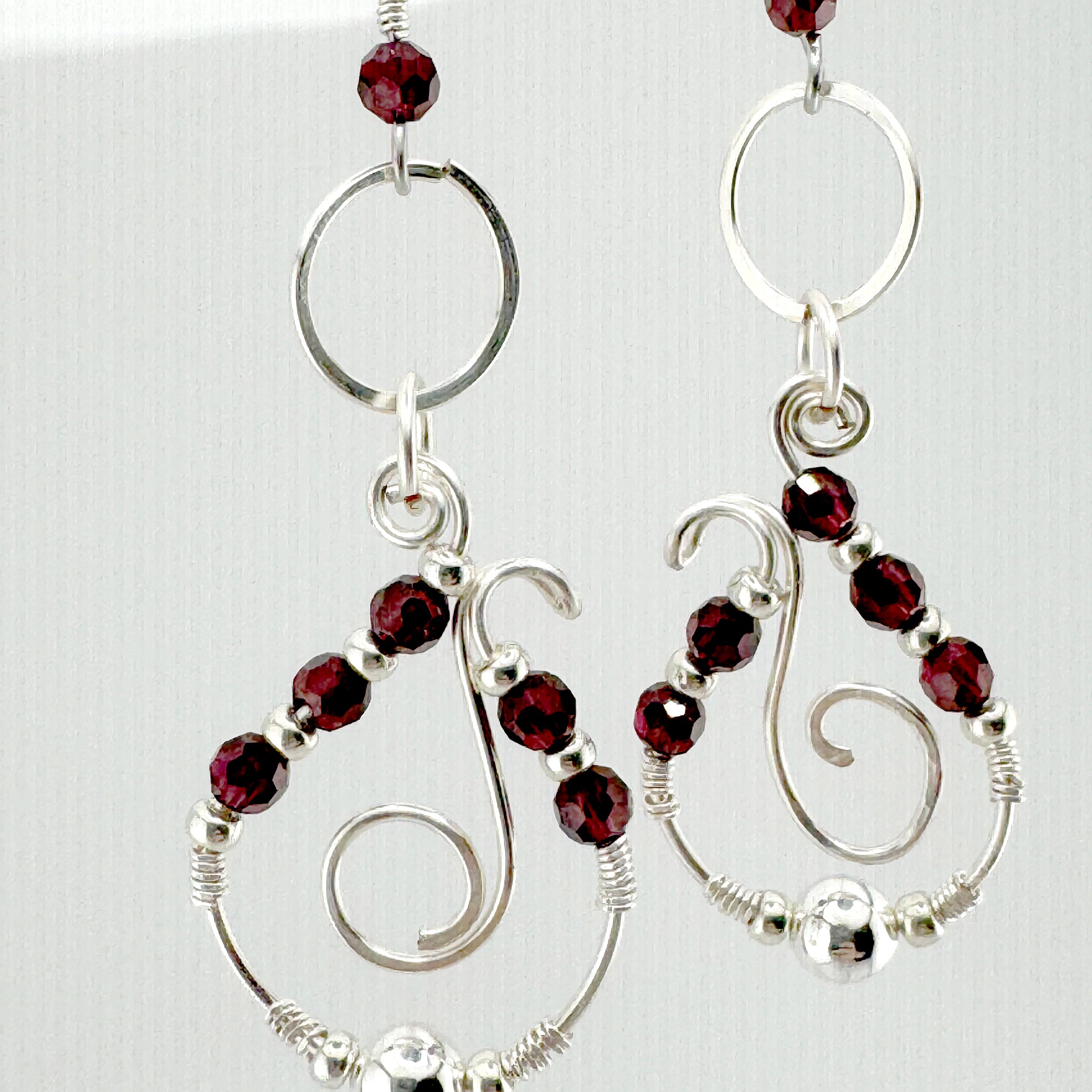 A pair of silver-toned dangle earrings with faceted dark red garnet stones, open circular hoops, and spiral wire details, shown against a light background.