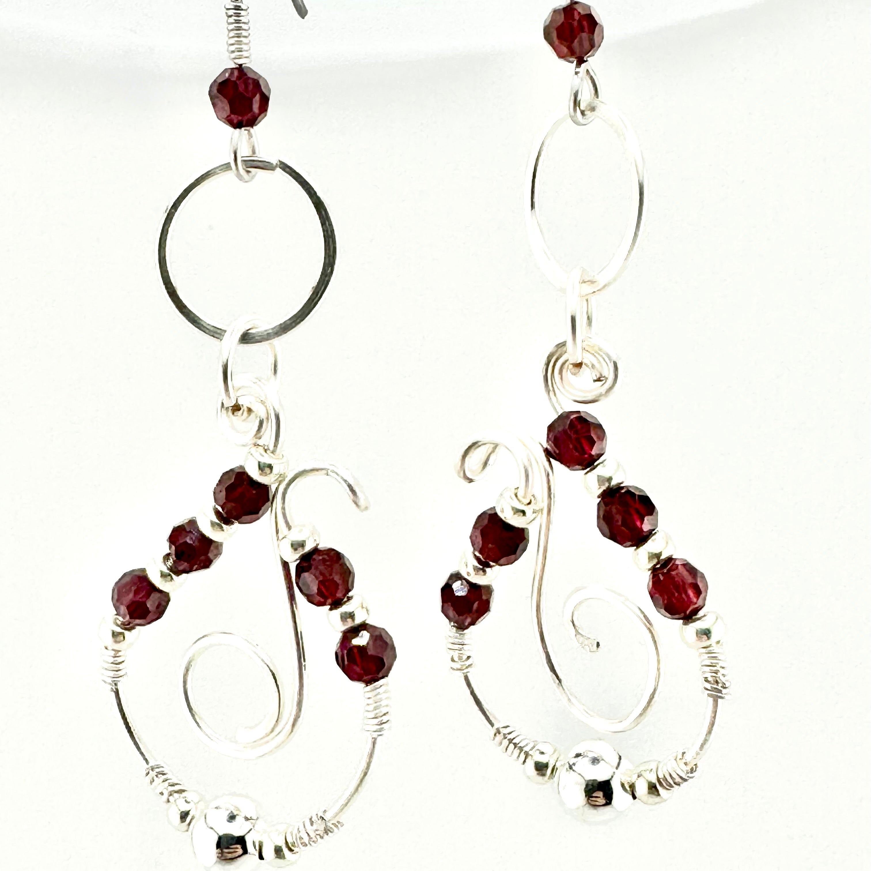 A pair of silver-toned dangle earrings with faceted dark red garnets, featuring circular and spiral wire designs on a white background.