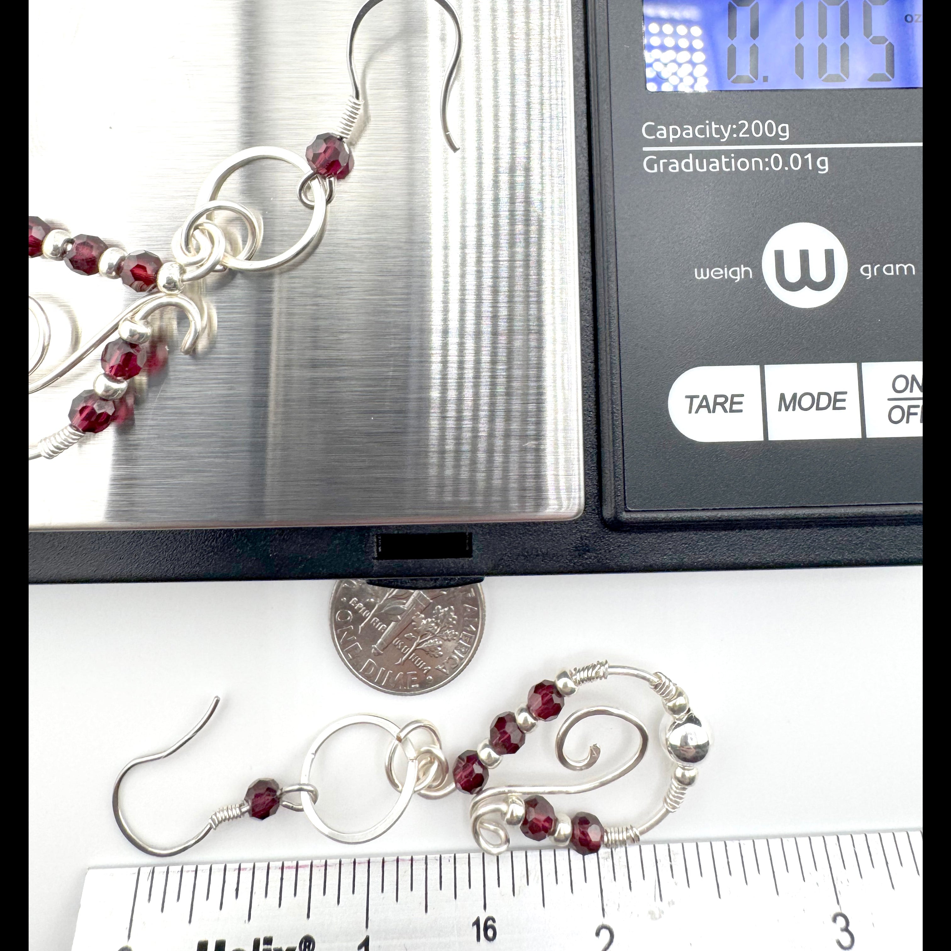 Silver-toned spiral drop earrings with red faceted garnets, shown on a digital scale alongside a dime and ruler for scale.