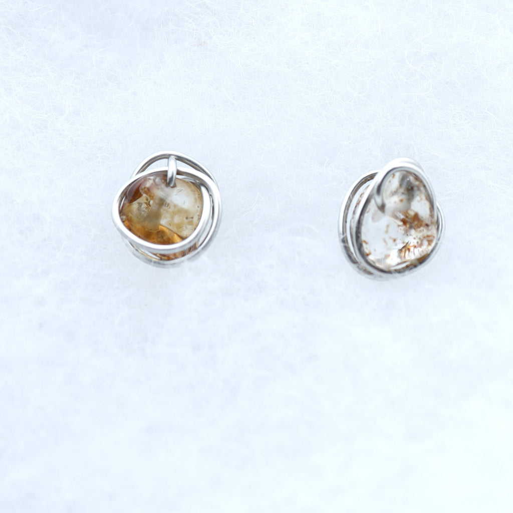 A pair of small silver stud earrings with smooth, organic-shaped quartz stones with warm golden-brown natural inclusions, displayed on a soft white textured fabric background.