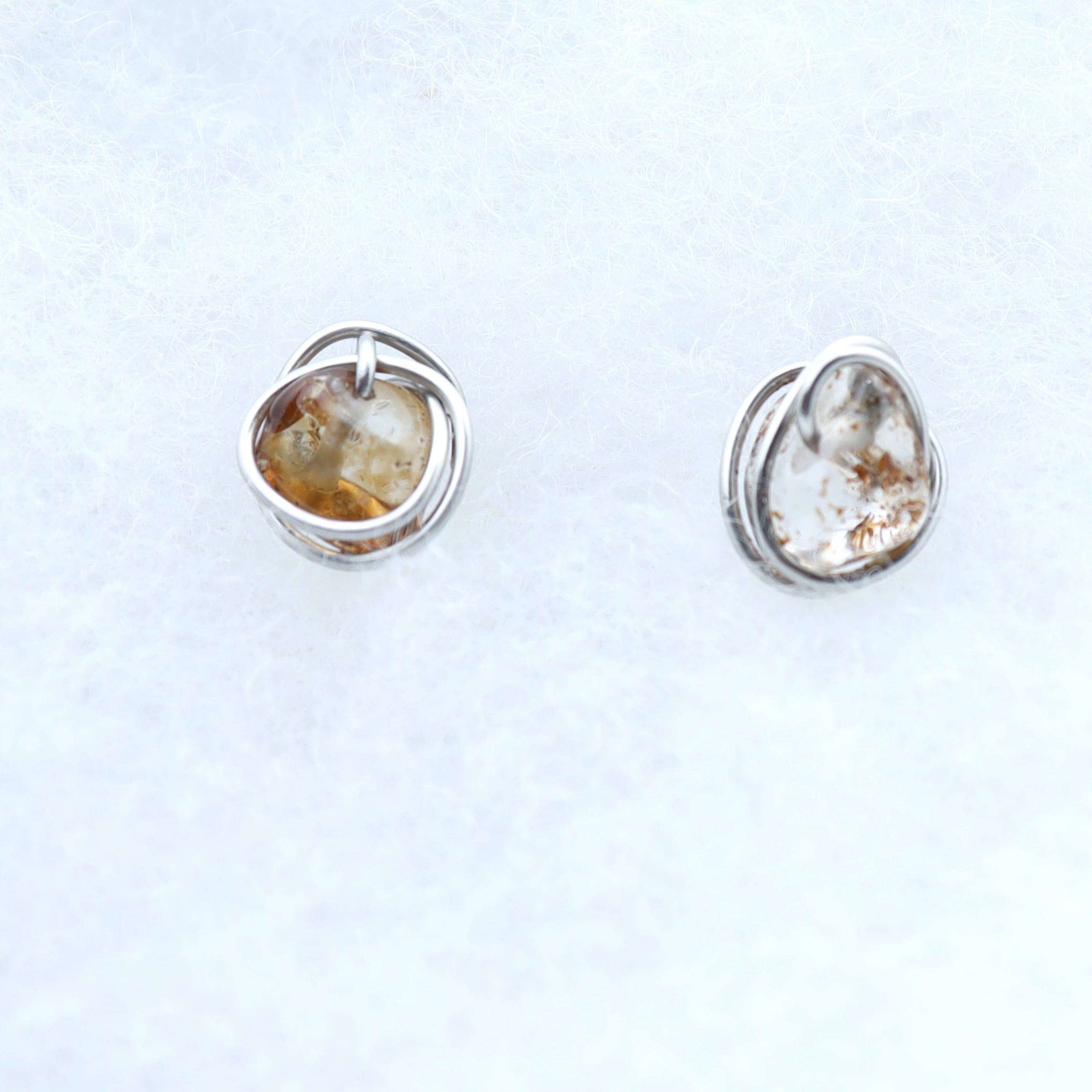 A pair of small silver stud earrings with smooth, organic-shaped quartz stones with warm golden-brown natural inclusions, displayed on a soft white textured fabric background.