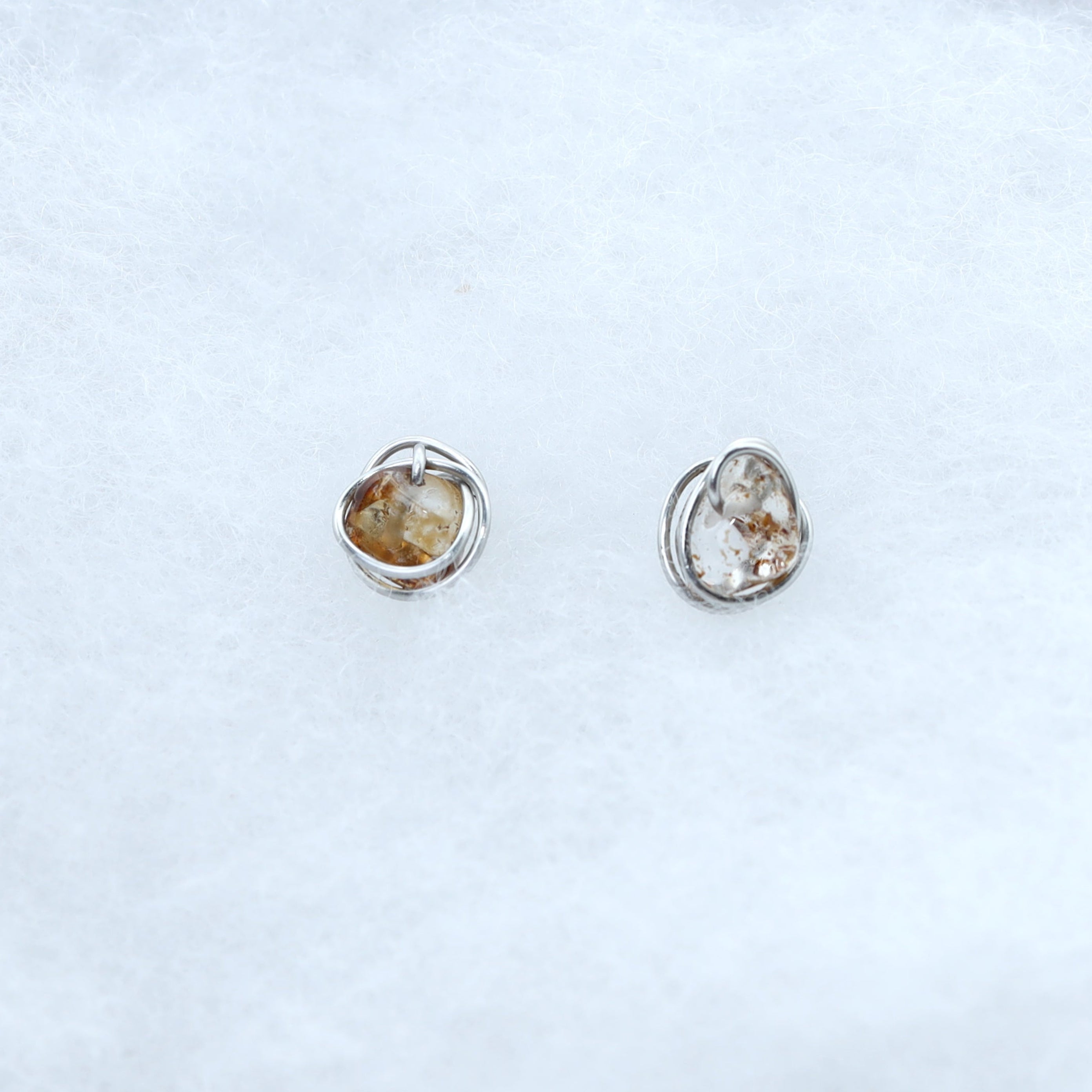 A pair of small silver-toned stud earrings with organic-shaped quartz stones with warm golden-brown natural inclusions
, displayed on a white textured jewelry box.