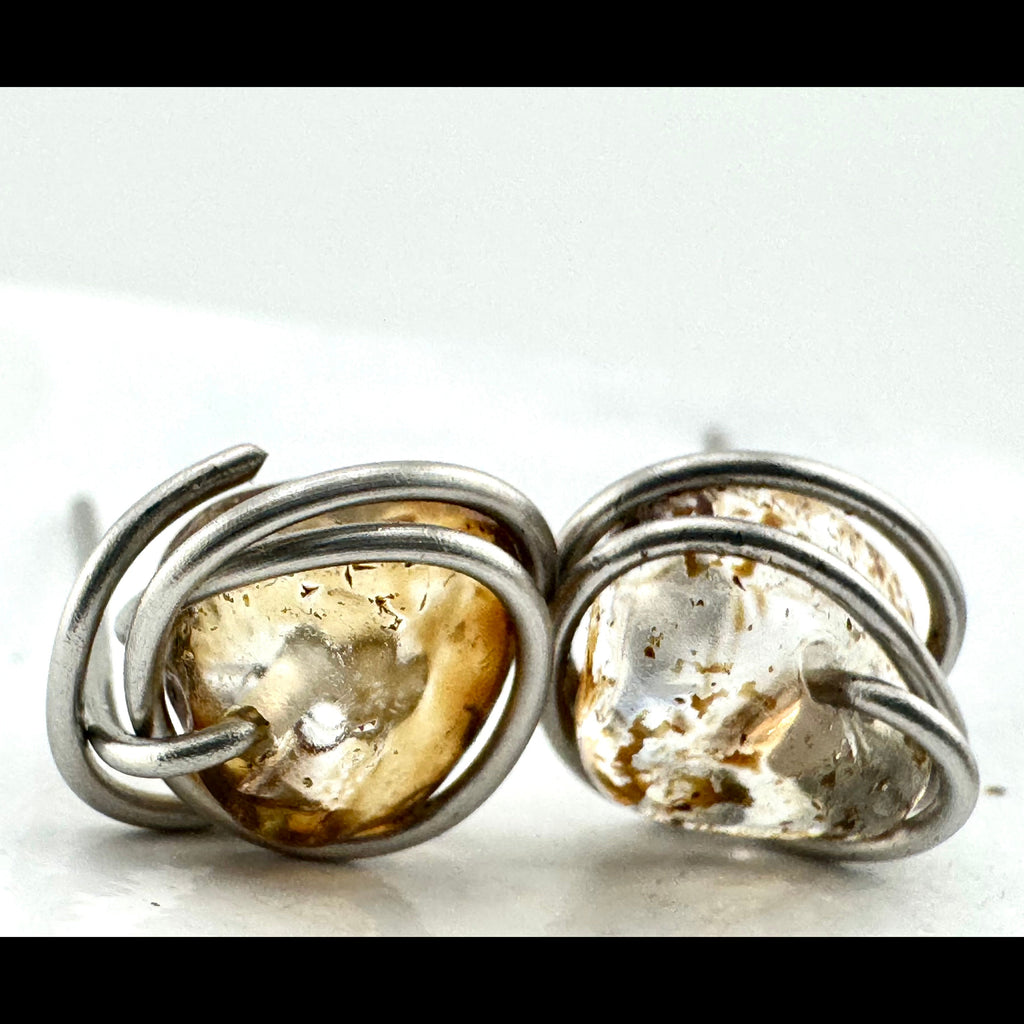 Close-up of a pair of handmade stud earrings featuring irregular quartz stones with warm golden-brown natural inclusions wrapped in stainless steel, shown on a plain white background.