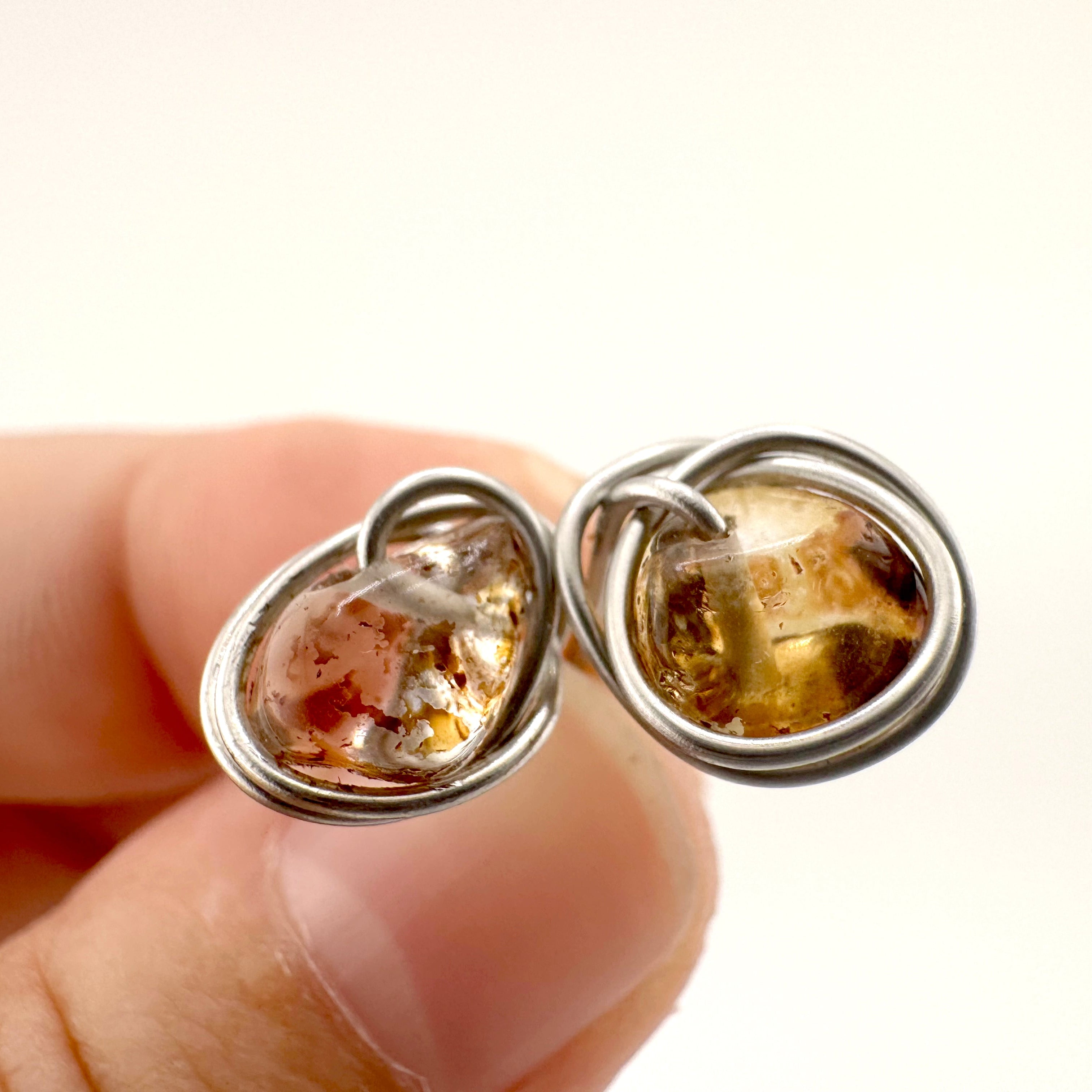 A close-up of fingers holding a pair of stainless steel wire-wrapped quartz stones with warm golden-brown natural inclusions stud earrings, against a plain white background.