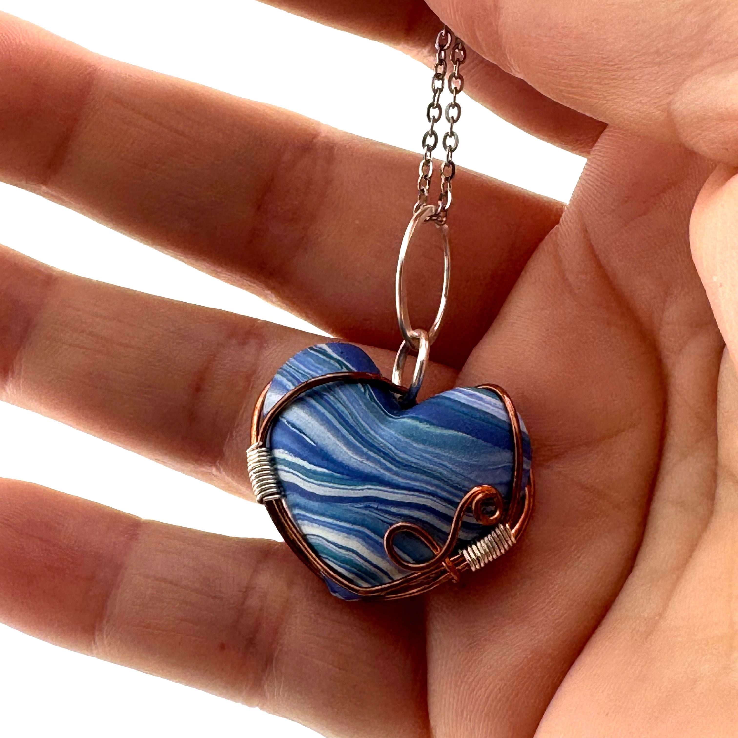 A heart-shaped pendant necklace with blue and white marbled clay, wrapped in copper and silver wire, held in the weaver's hand against a white background.