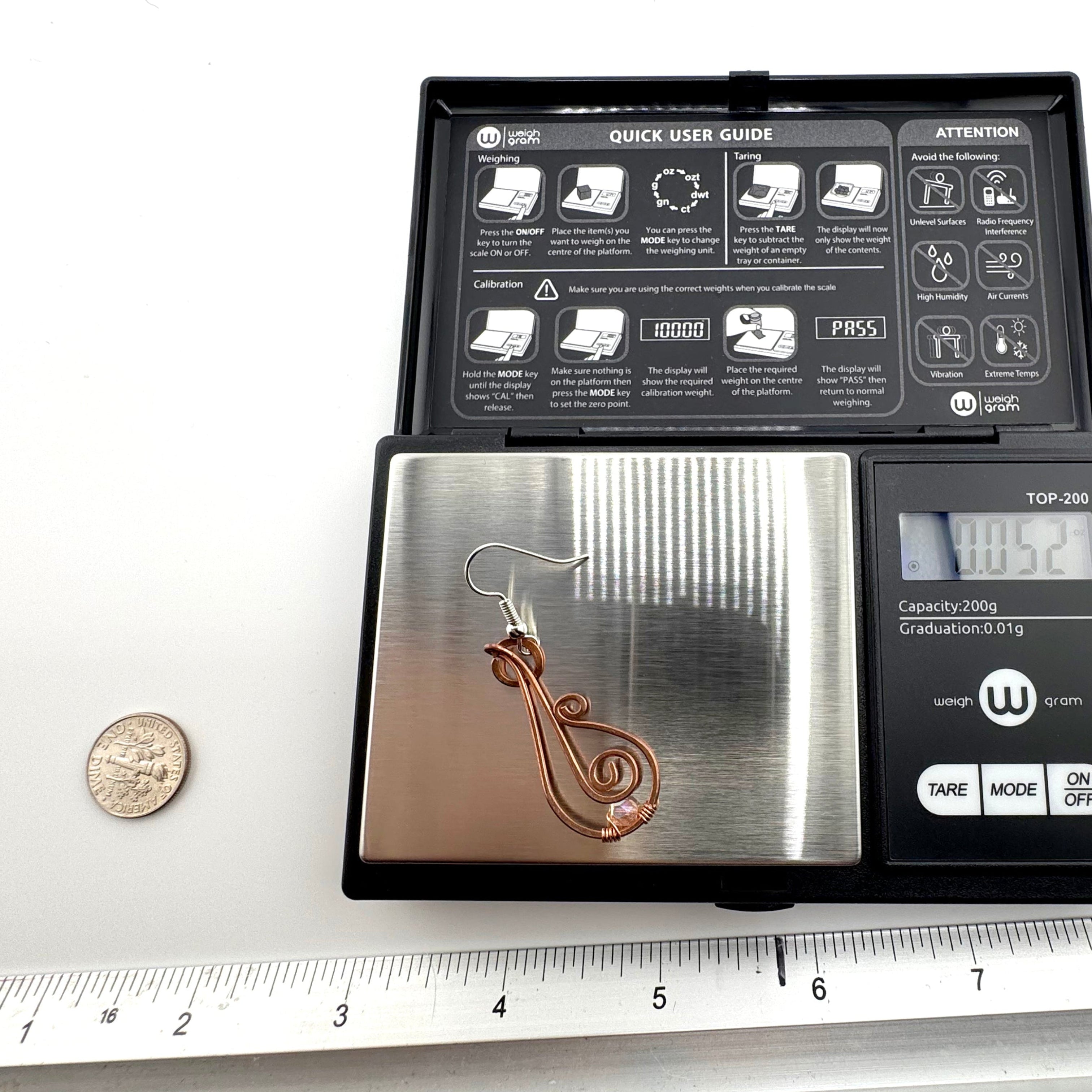 A small digital pocket scale with a metallic weighing platform is shown open, displaying a copper-colored the earring on its platform. The scale is placed on a white surface with a coin and a metal ruler nearby for size reference.