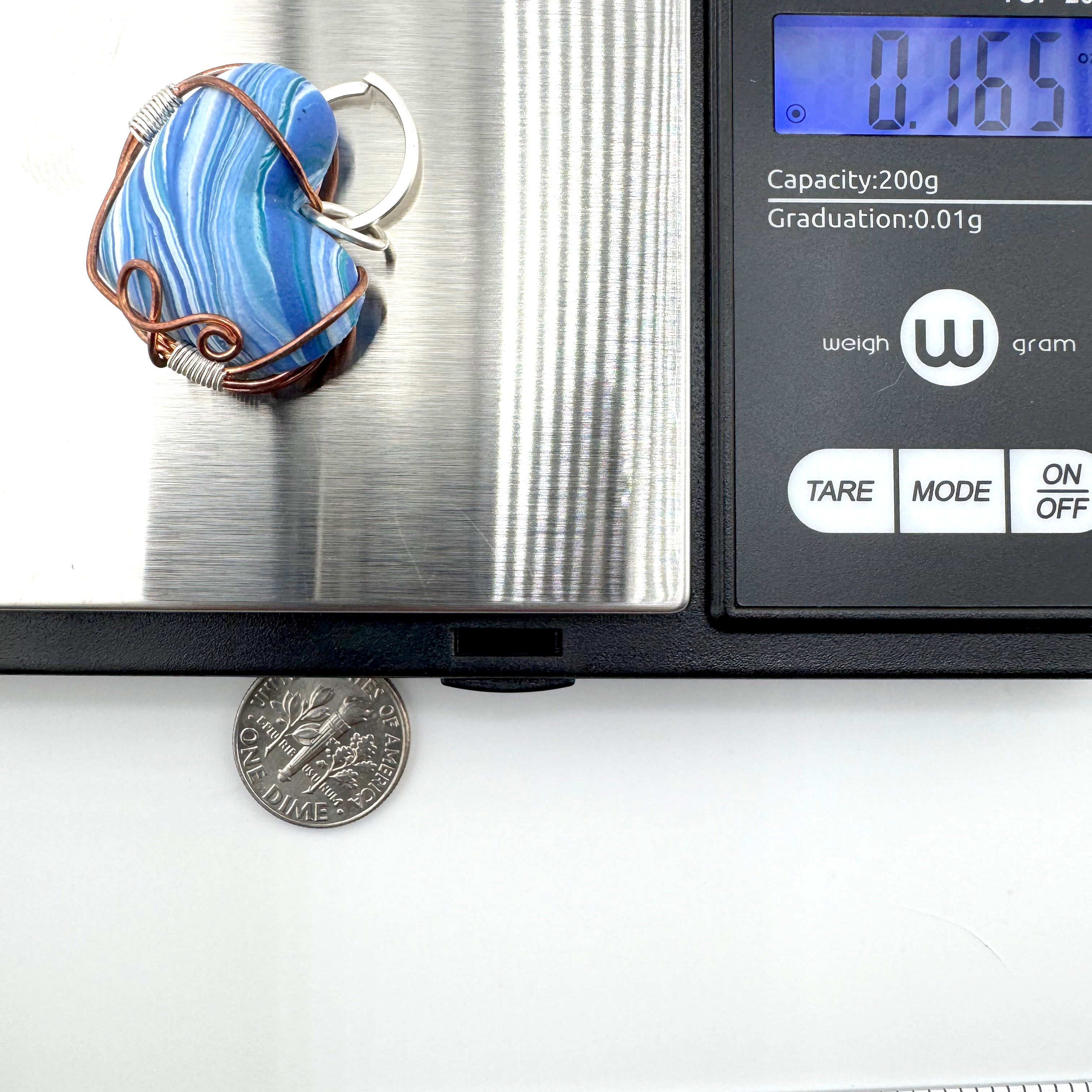 A blue and white marbled clay pendant with copper wire wrapping and a silver metal loop, shown on a digital scale next to a coin for scale.