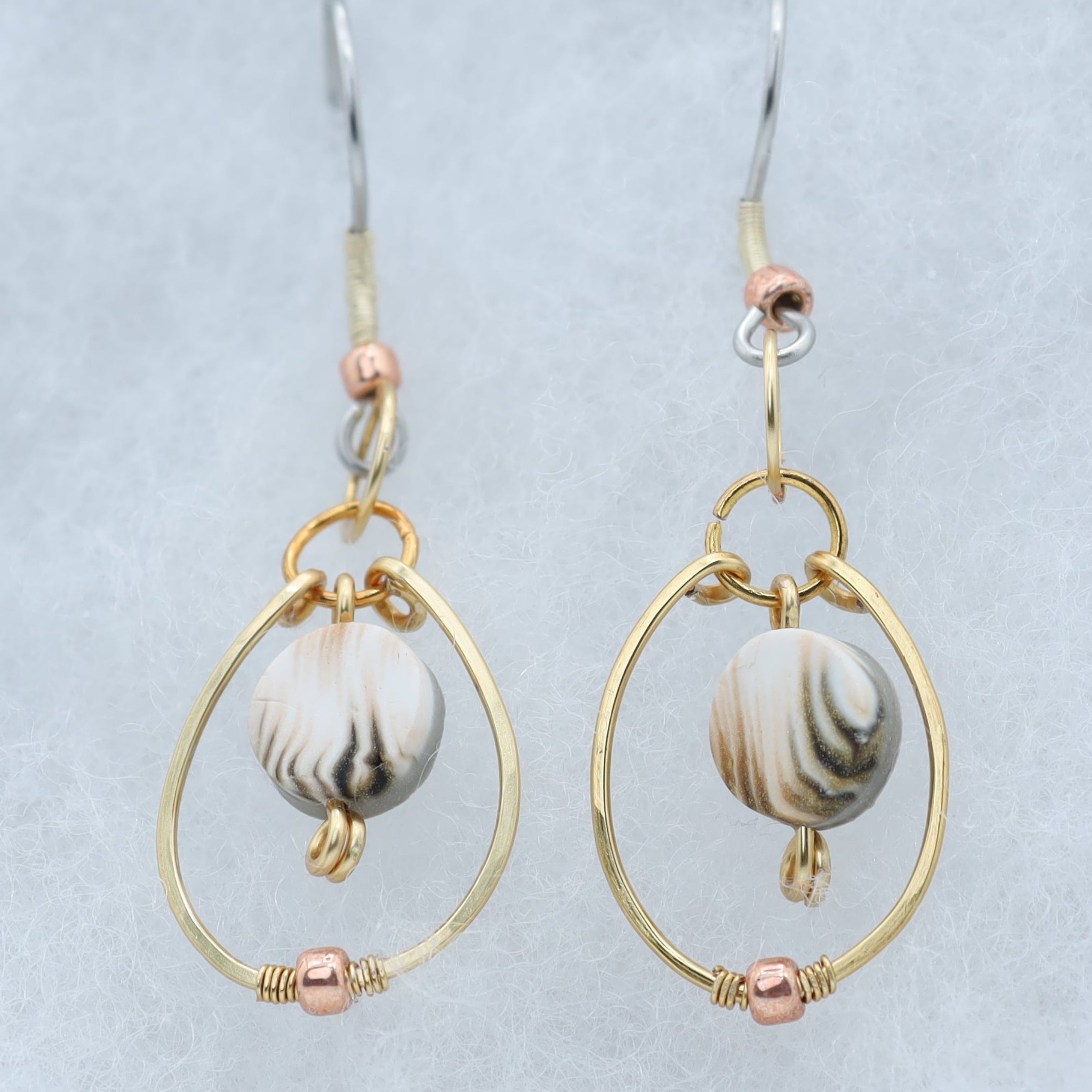 A pair of gold and rose gold drop earrings with white, tan and gray marbled clay beads, displayed on a light gray velvet jewelry box.