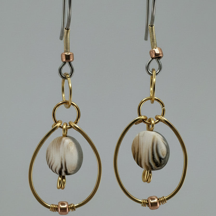 A pair of gold and silver drop earrings with circular wire frames and a central round  clay bead featuring natural black and cream banding, displayed against a neutral gray background.