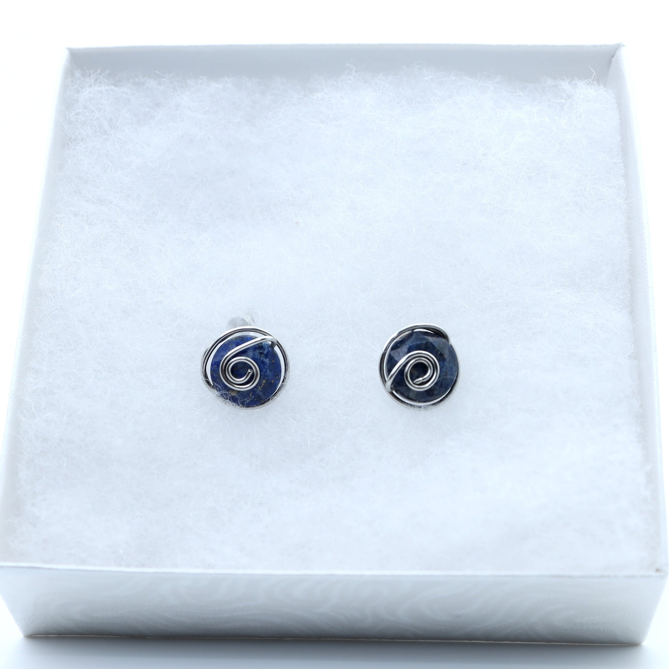 A pair of small  stud earrings featuring dark blue aventurine 10mm faceted stones held in hand-shaped stainless steel spiral wire details,, displayed on a white textured insert inside a white gift box.