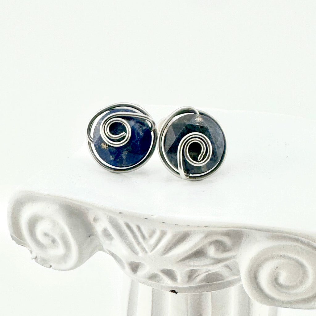 A pair of spiral stud earrings featuring dark blue aventurine 10mm faceted stones held in hand-shaped stainless steel spiral wire details,, displayed on a white decorative column against a white background.