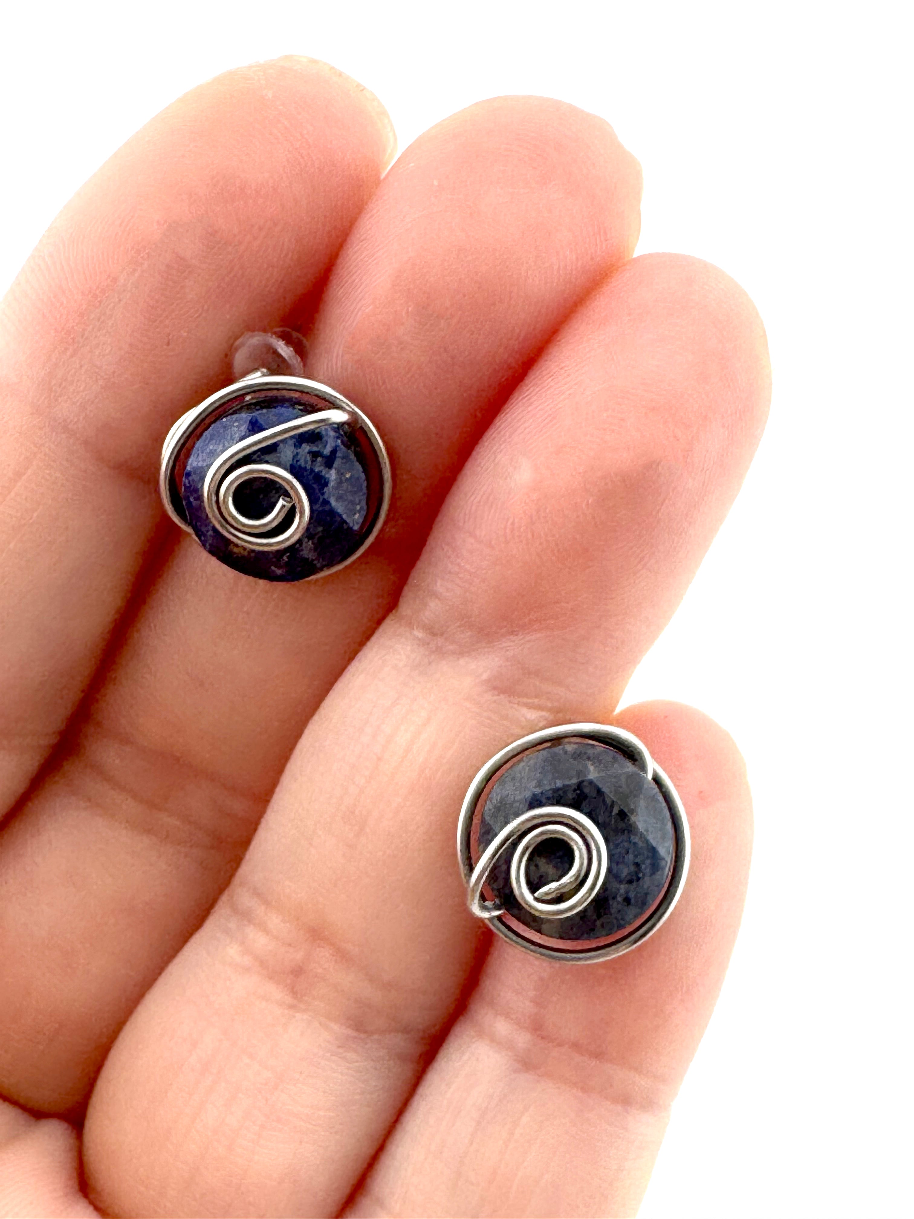 A close-up of a hand holding two small  stud earrings with dark blue aventurine 10mm faceted stones held in hand-shaped stainless steel spiral wire details against a white background.