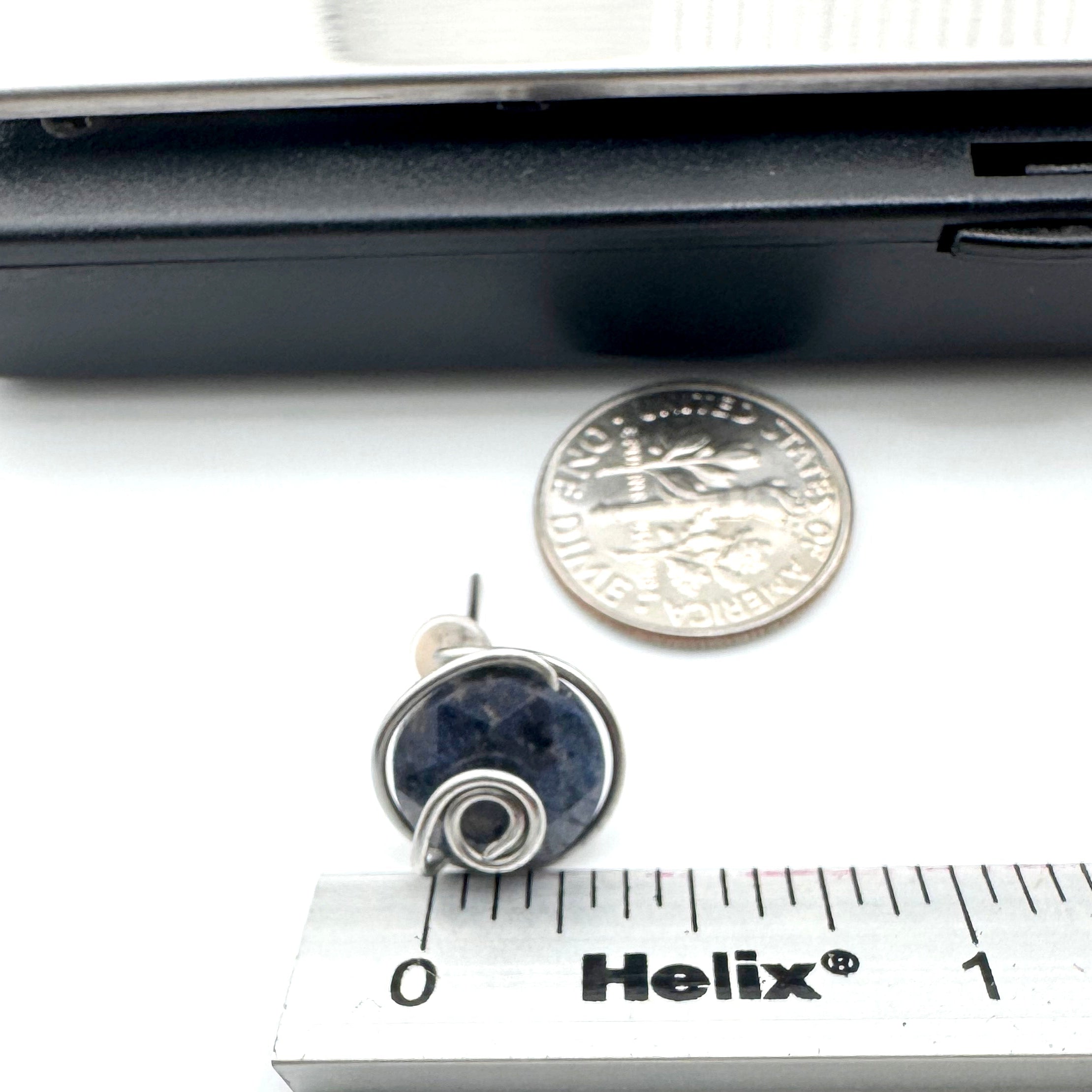 A small  stud earring  featuring dark blue aventurine 10mm faceted stones held in hand-shaped stainless steel spiral wire details, shown next to a dime and a Helix ruler for scale on a white background.