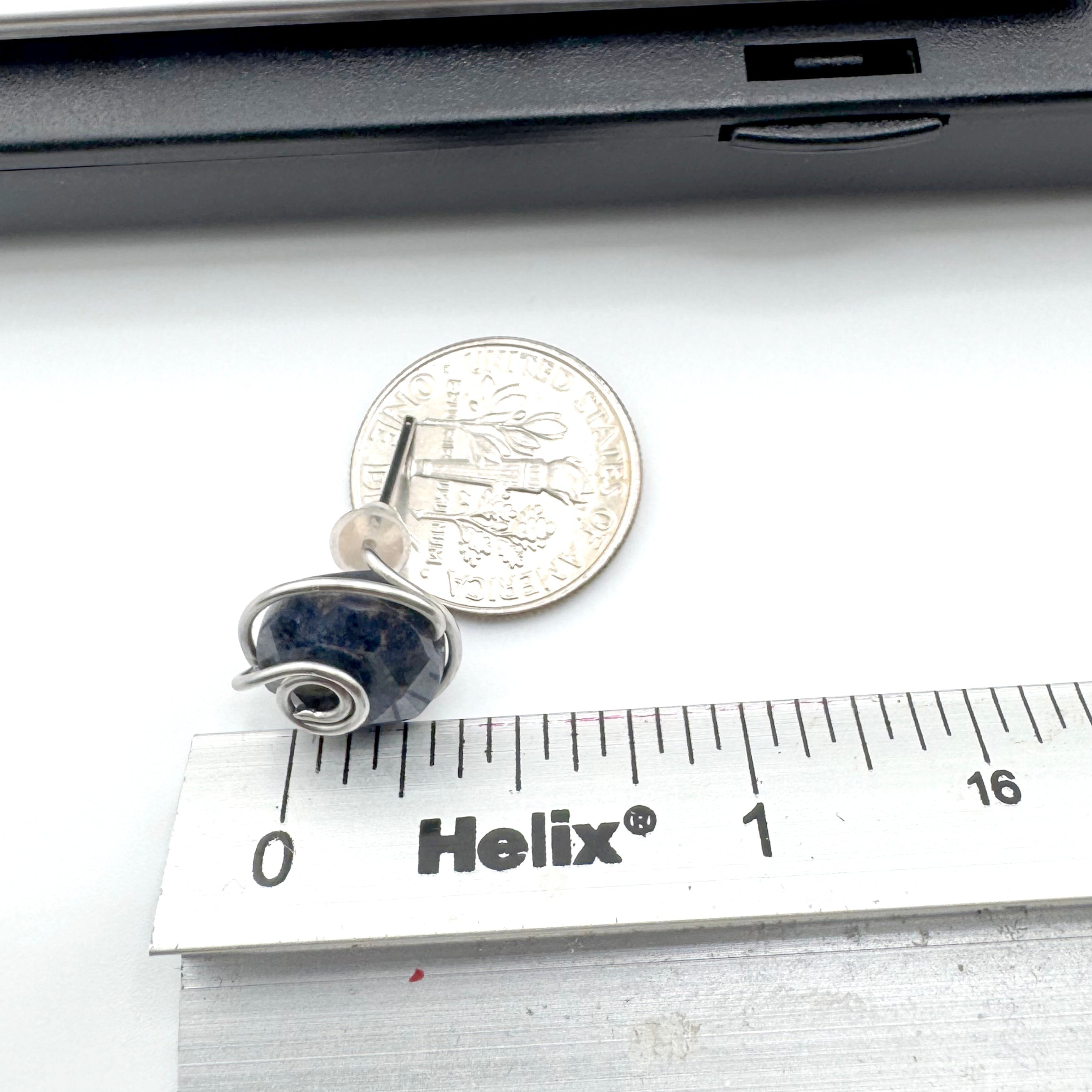 A pair of  wire-wrapped earrings featuring dark blue aventurine 10mm faceted stones held in hand-shaped stainless steel spiral wire details, shown next to a US quarter for scale and a Helix ruler on a white surface.