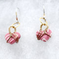 A pair of handmade dangle earrings featuring pink and white marbled heart-shaped clay beads wrapped in gold-toned wire, hanging from silver fishhook ear wires on a textured white background.