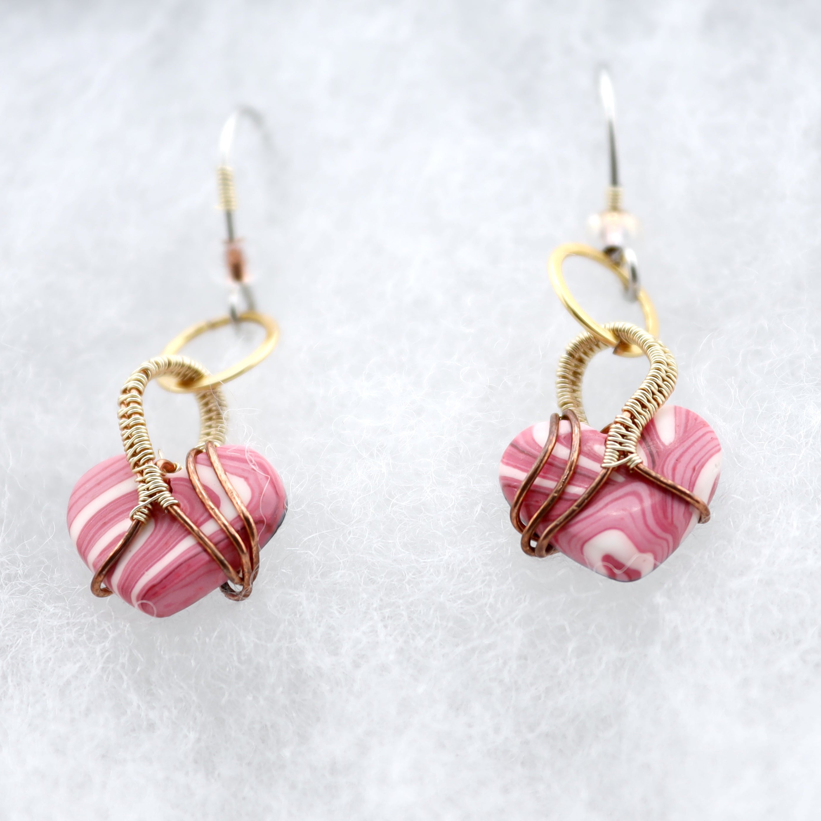 A pair of handmade dangle earrings featuring pink and white marbled heart-shaped clay beads wrapped in gold-toned wire, hanging from silver fishhook ear wires on a textured white background.